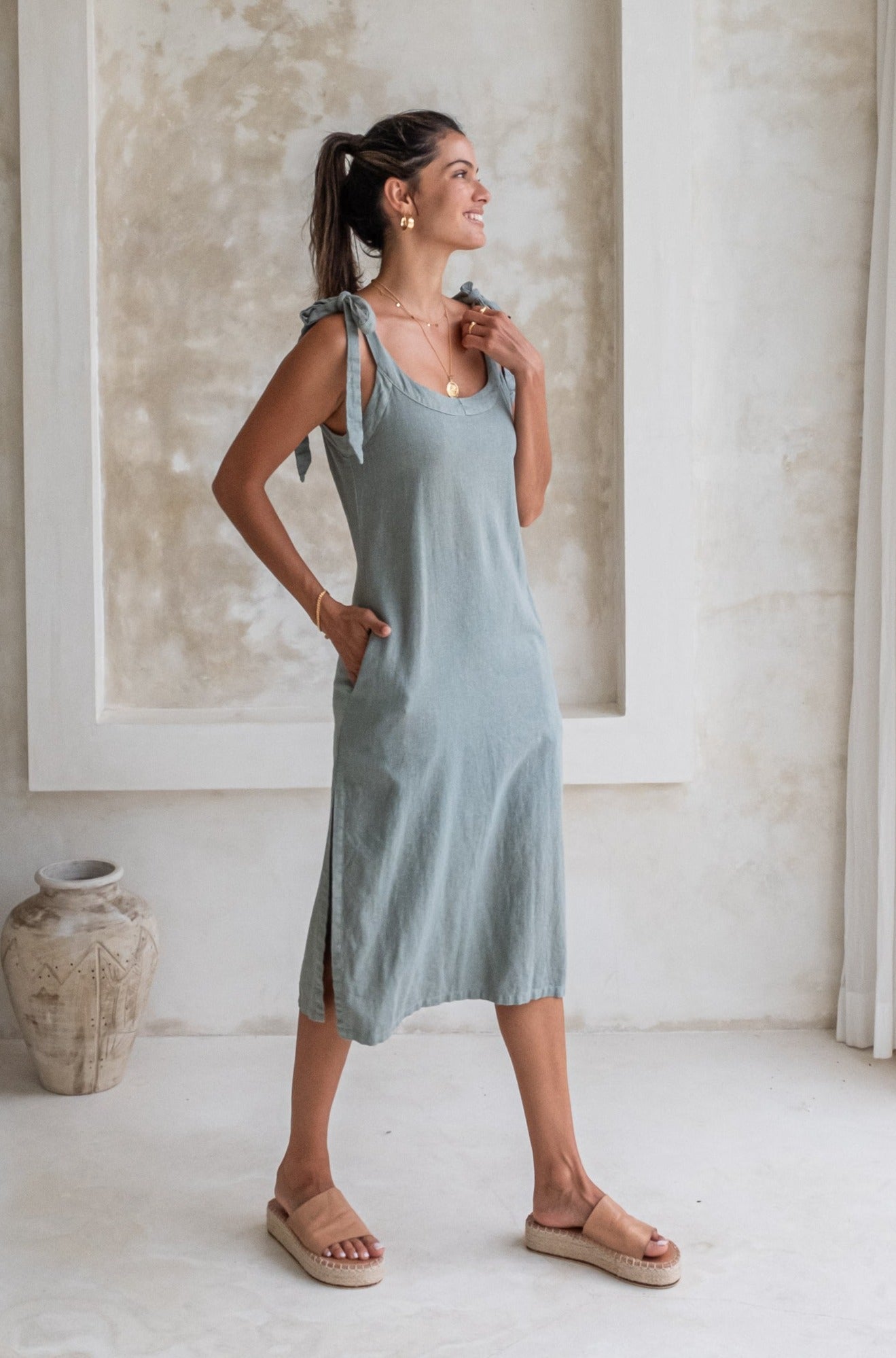 A woman in a light green midi dress with tied straps and beige espadrille sandals stands in a minimalist room, looking to the side with a smile.