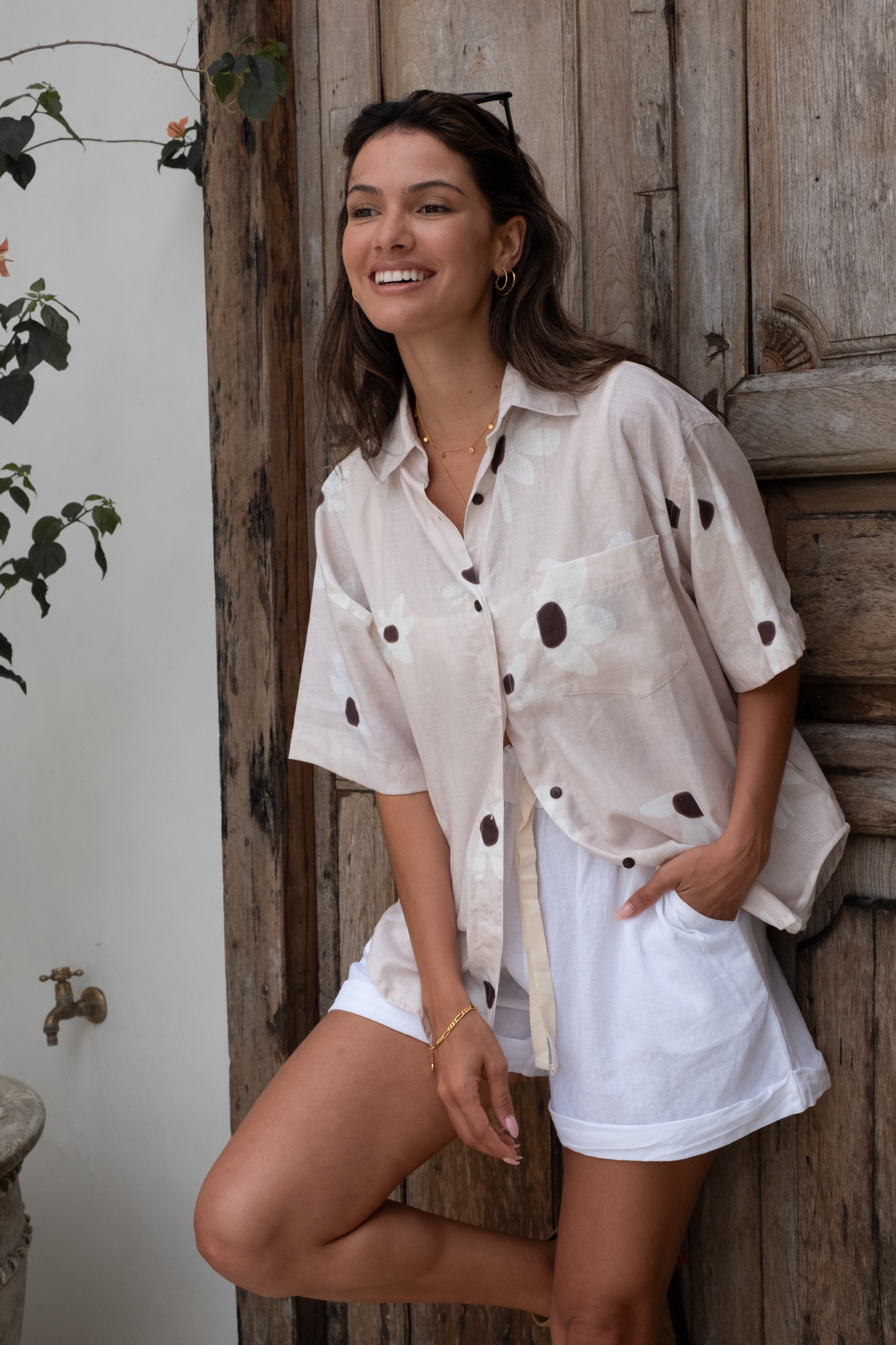 A woman wearing light pink floral pajama shorts and a matching loose shirt stands against a rustic wooden door