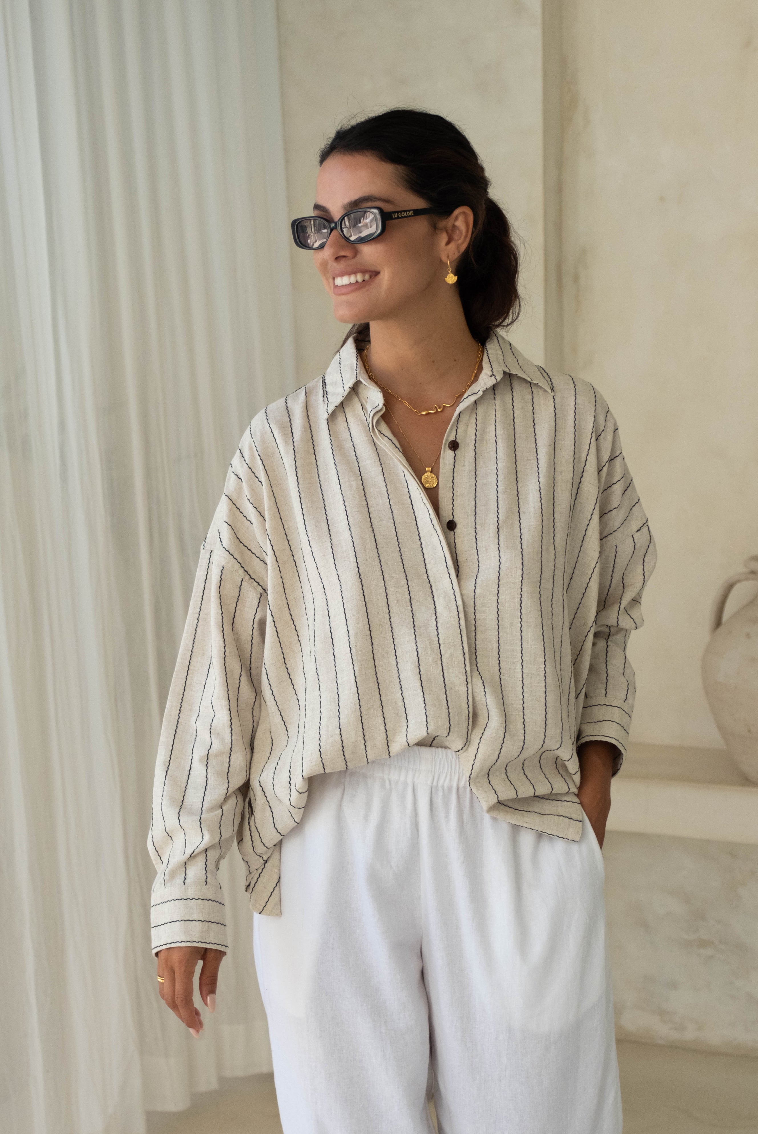 A person standing with a smile, hands in pockets, wearing sunglasses, a striped shirt, and white pants.
