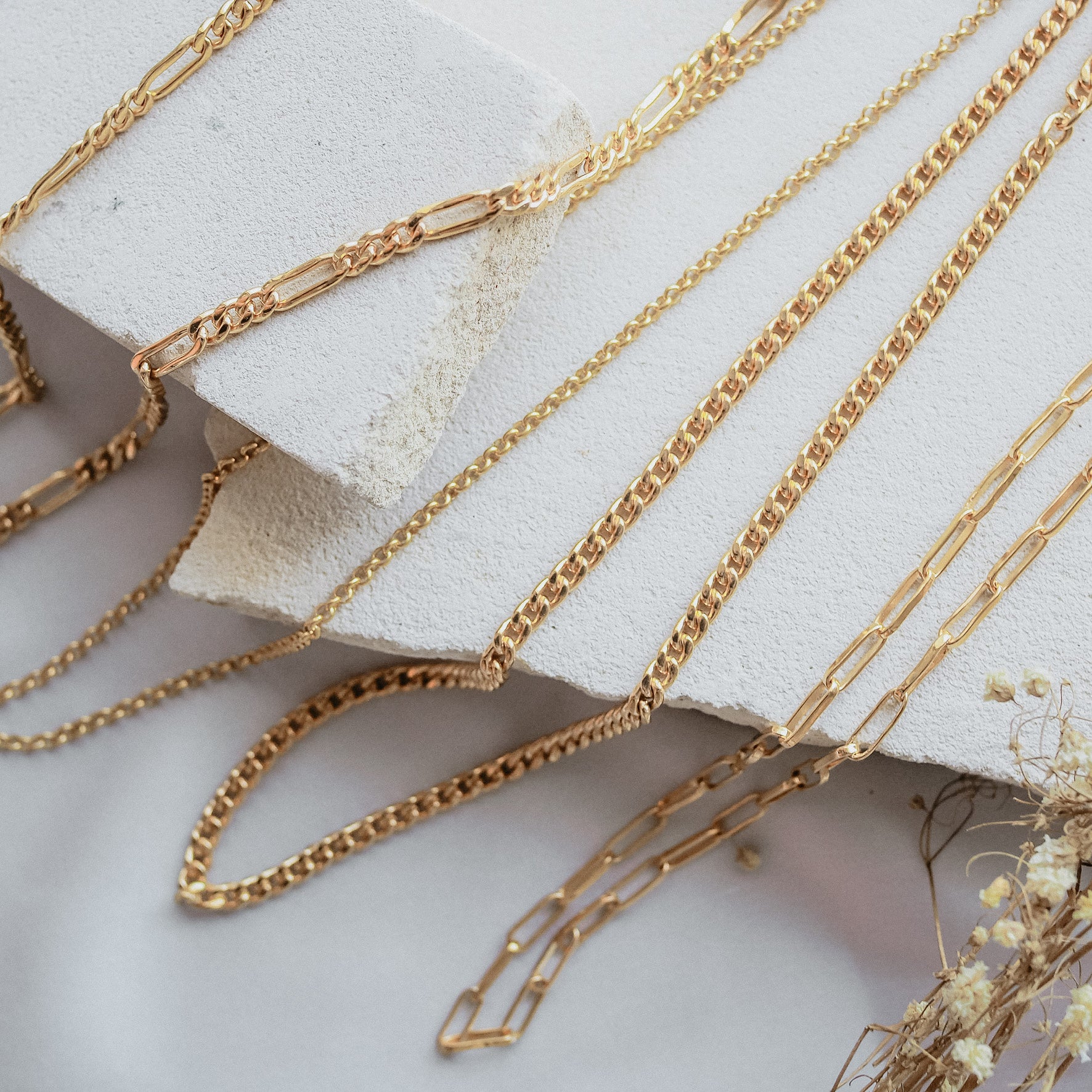 Multiple gold chain necklaces with different link styles displayed on a textured beige stone with dried flowers in the corner