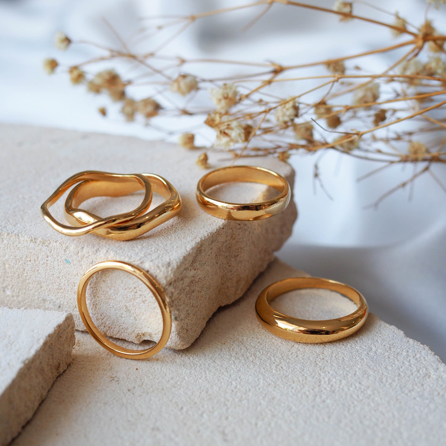 Gold rings, including wavy and smooth designs, displayed on textured beige stones with dried flowers.