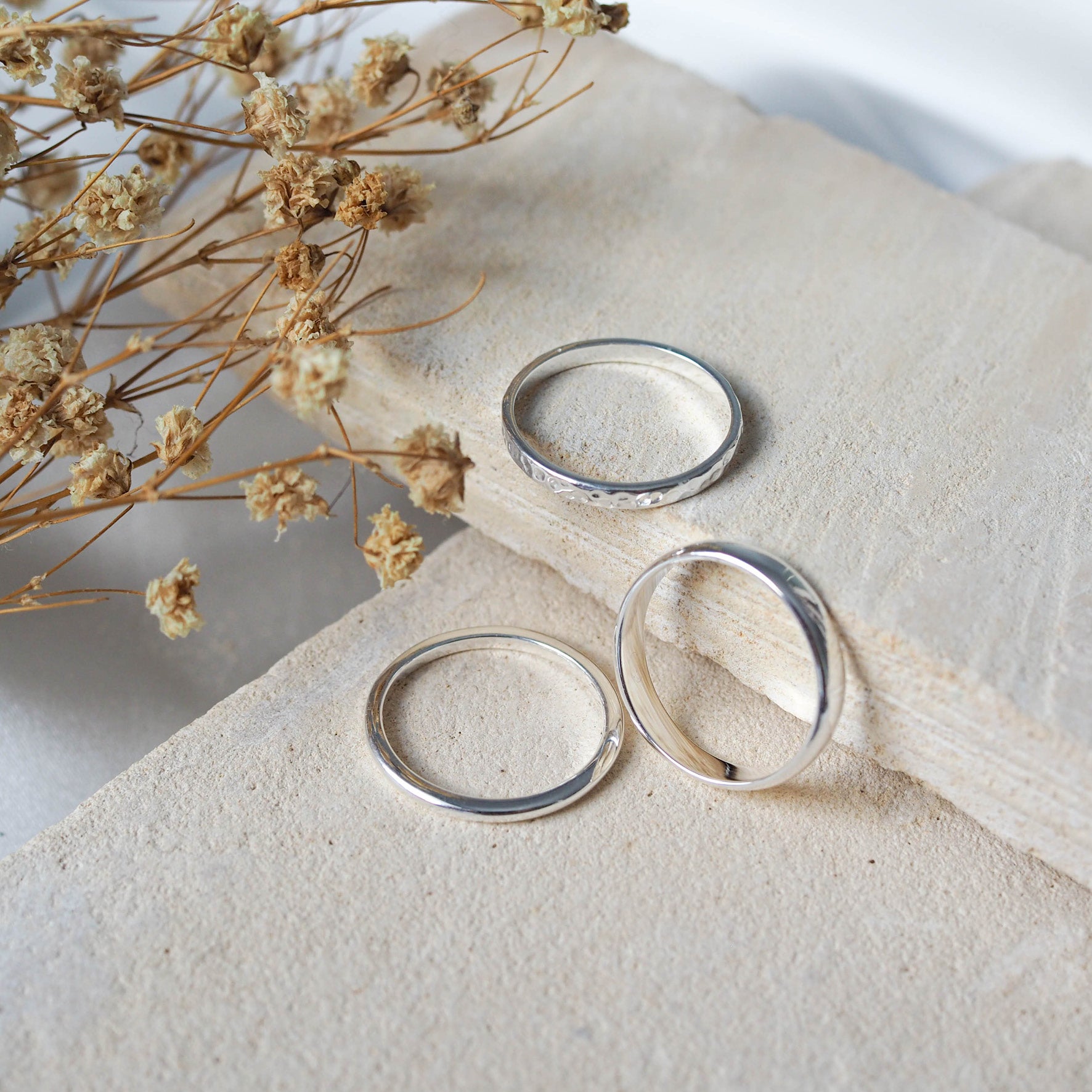 Silver rings, including wavy and smooth designs, displayed on textured beige stones with dried flowers.