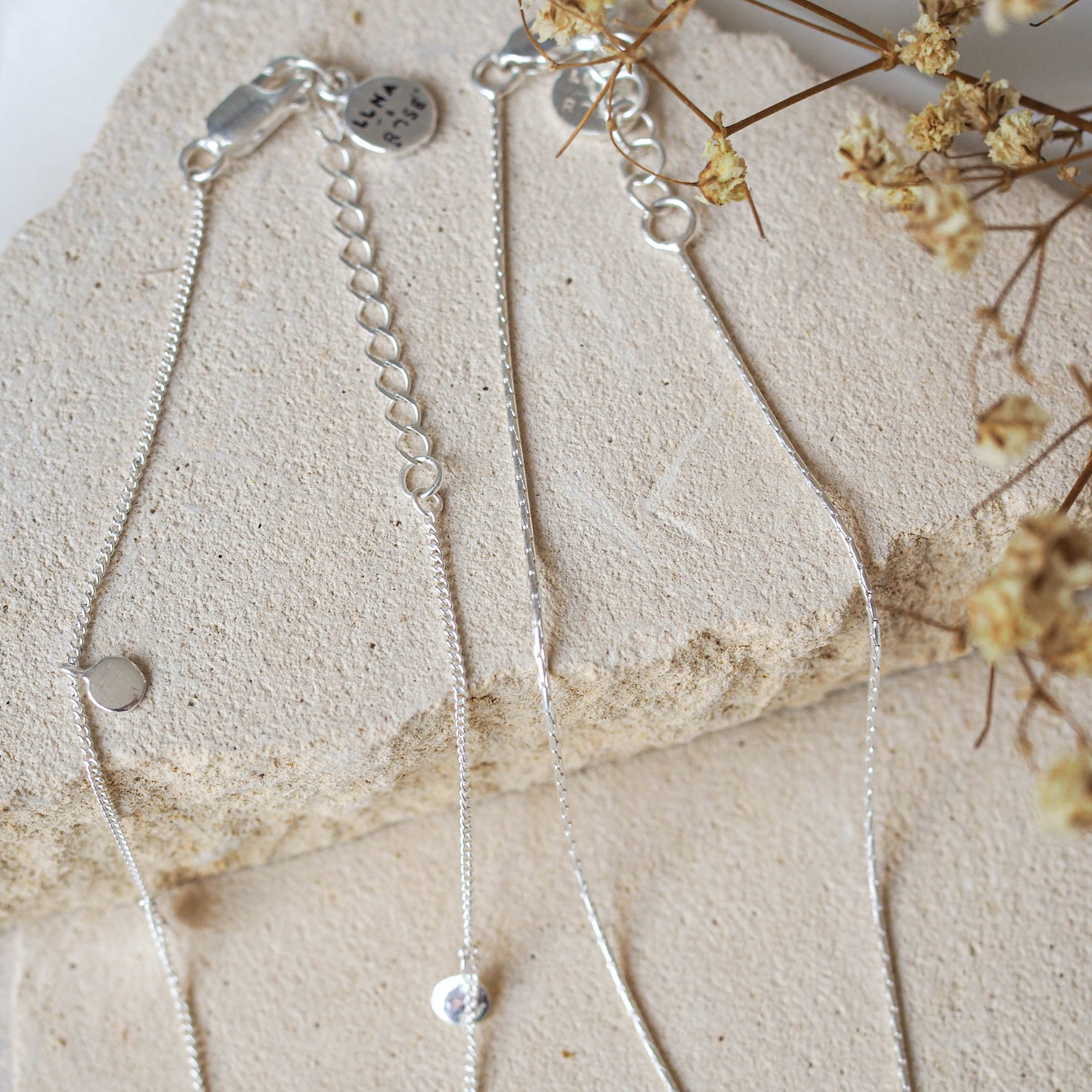 Two silver anklets, one with elongated links and the other a delicate chain, displayed on beige stone with dried flowers.