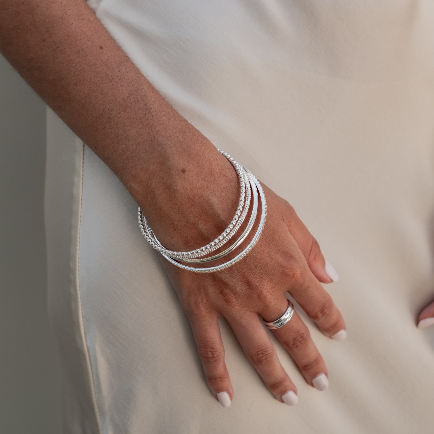 Multiple silver bracelets, including a beaded one, worn on both wrists along with a silver ring on a finger, against a light background.