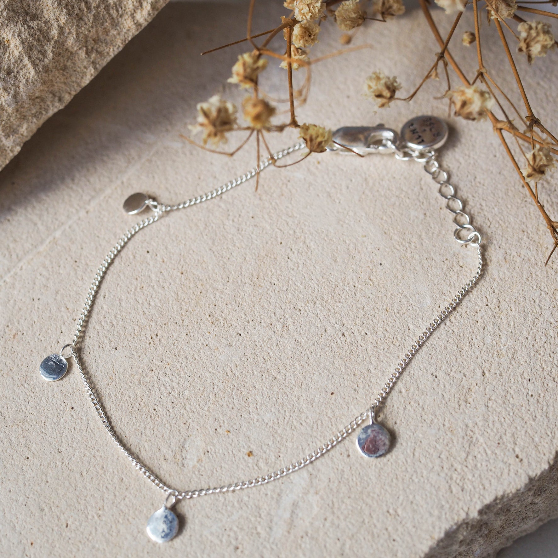 A delicate silver bracelet with small circular charms, displayed on beige stone with dried flowers.