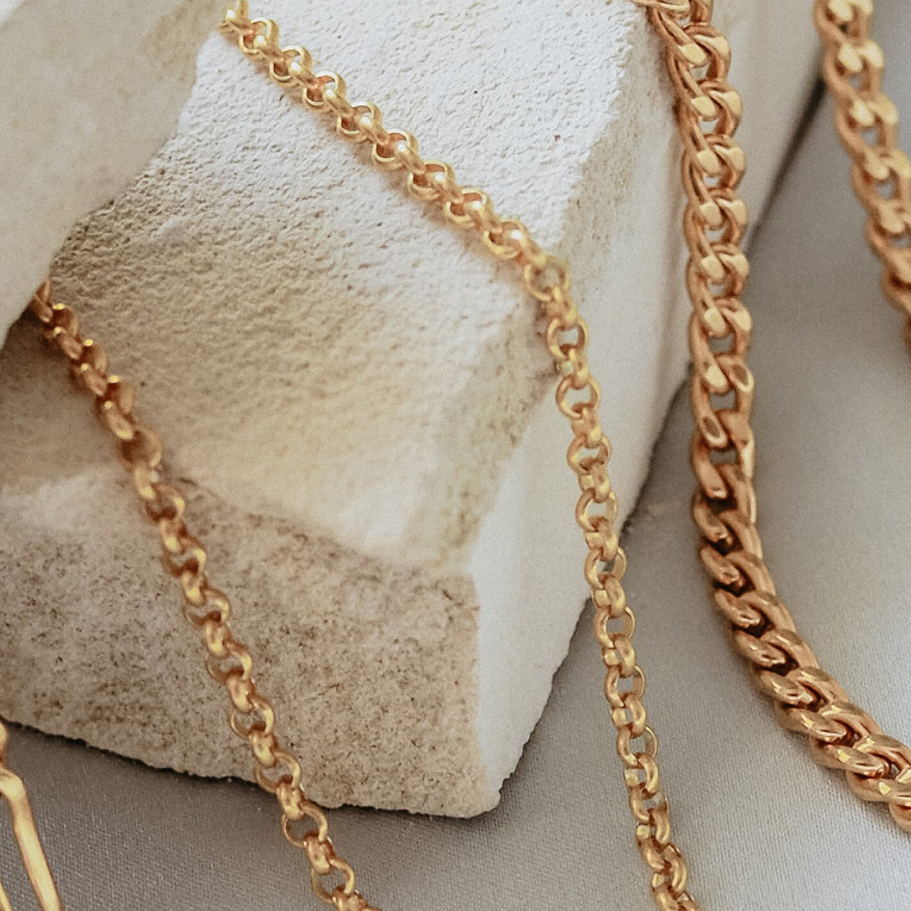 A gold chain bracelet with an adjustable clasp and circular charm, displayed on beige stone with dried flowers.