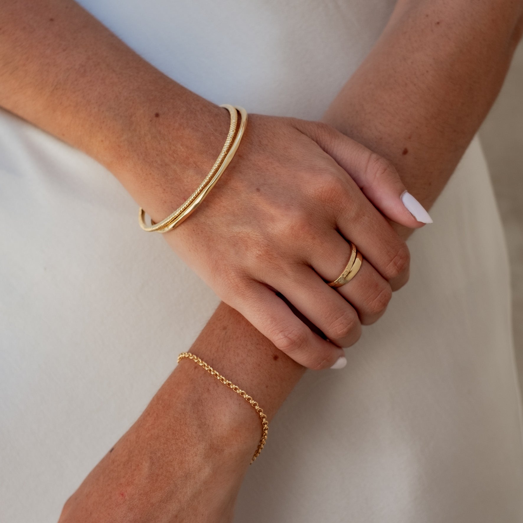 A woman's crossed arms, wearing a gold chain bracelet, a gold ring, and gold bangles, against a light-colored background.