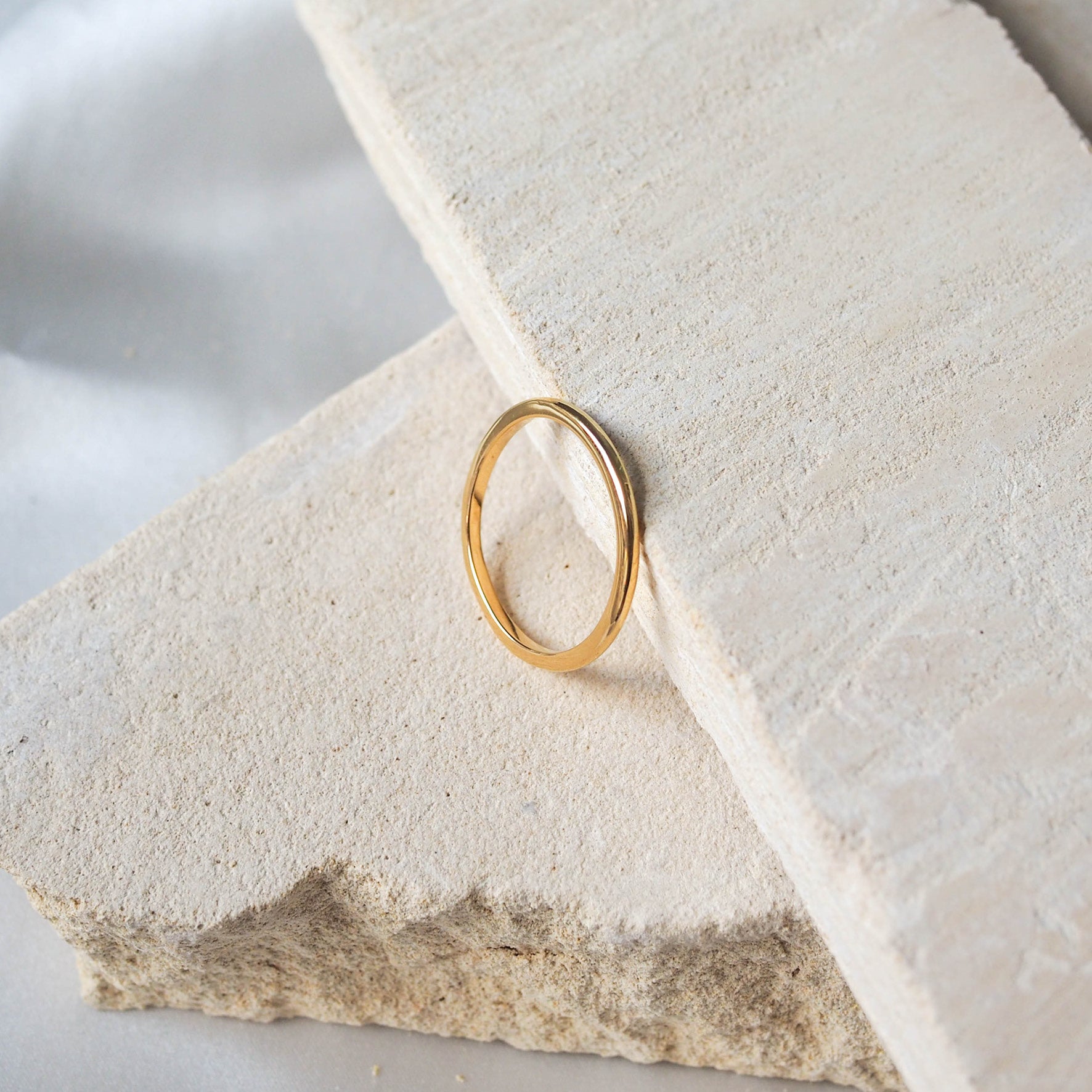 A single gold ring displayed on a textured beige stone, against a soft white background.