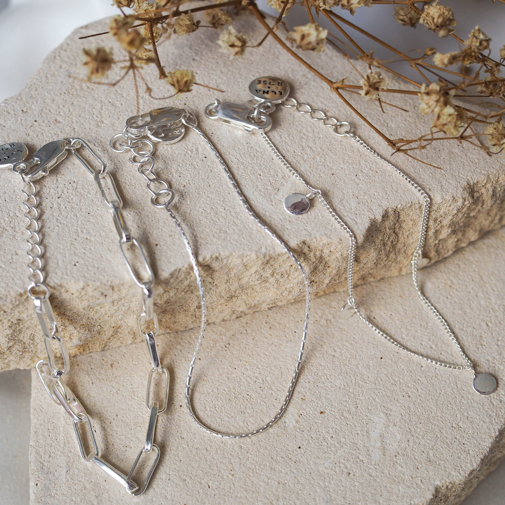 Three delicate silver bracelets with small circular charms and adjustable clasps, displayed on beige stone with dried flowers.