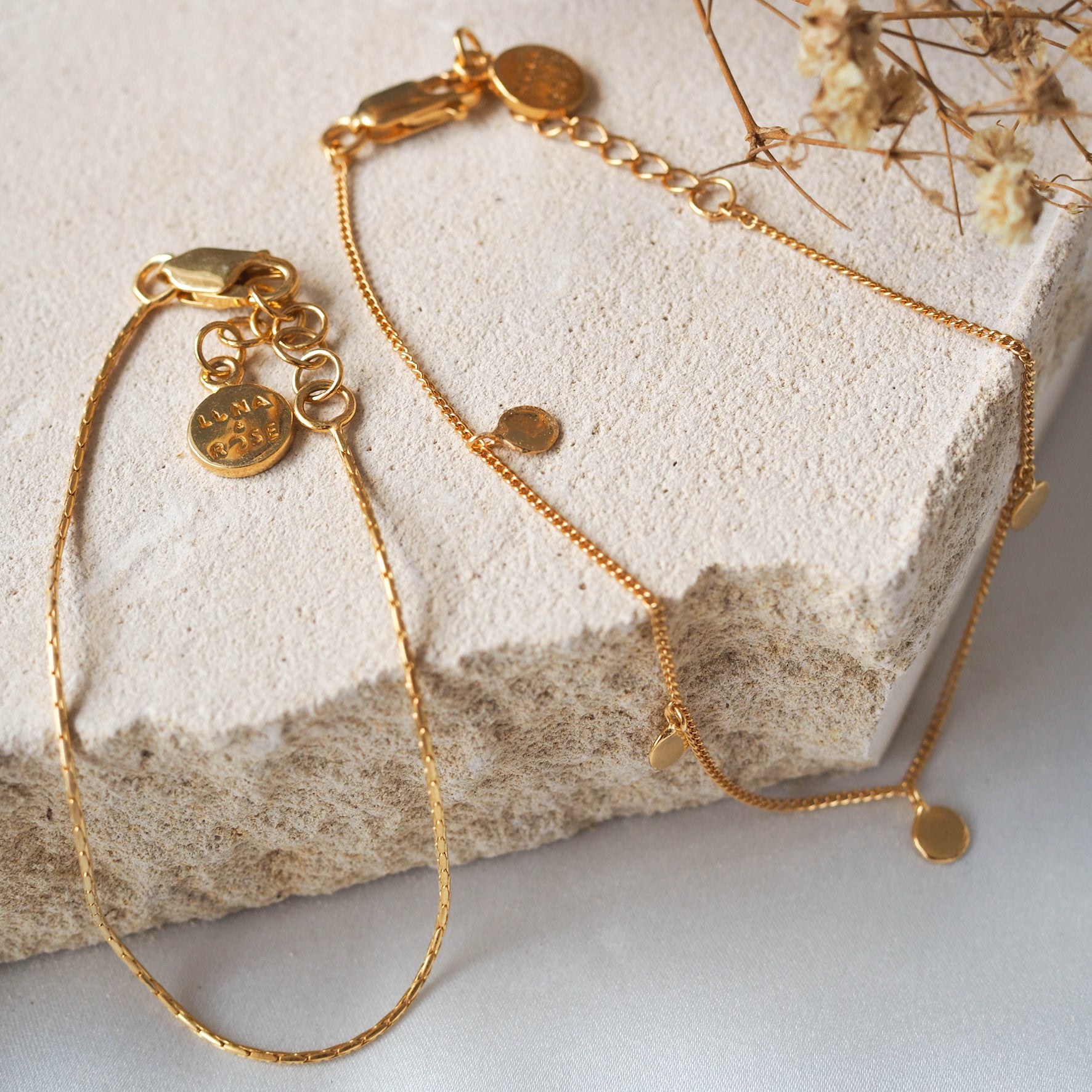 Two delicate gold bracelets with small circular charms and adjustable clasps, displayed on beige stone with dried flowers.