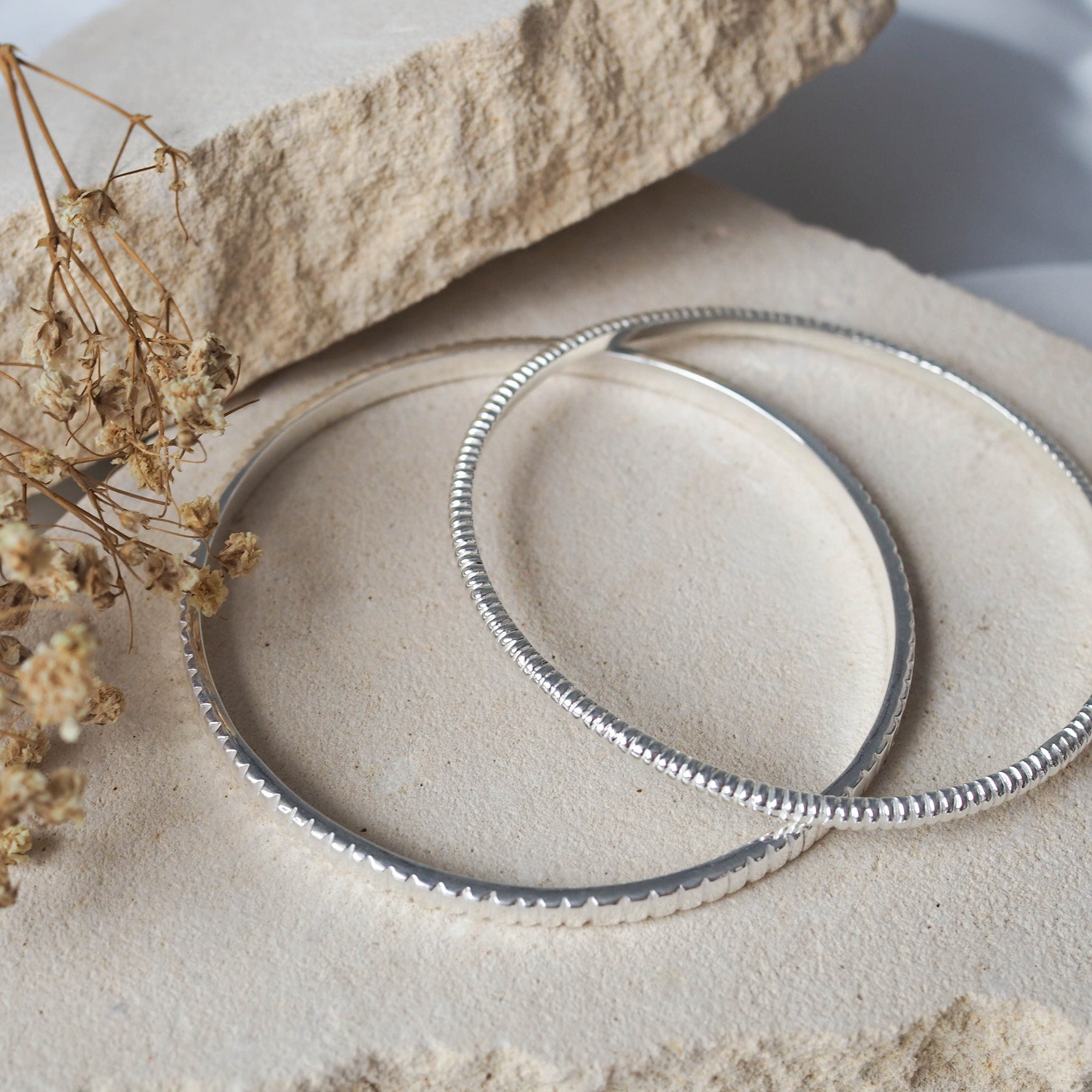 two bracelets silver displayed on beige stone with dried flowers