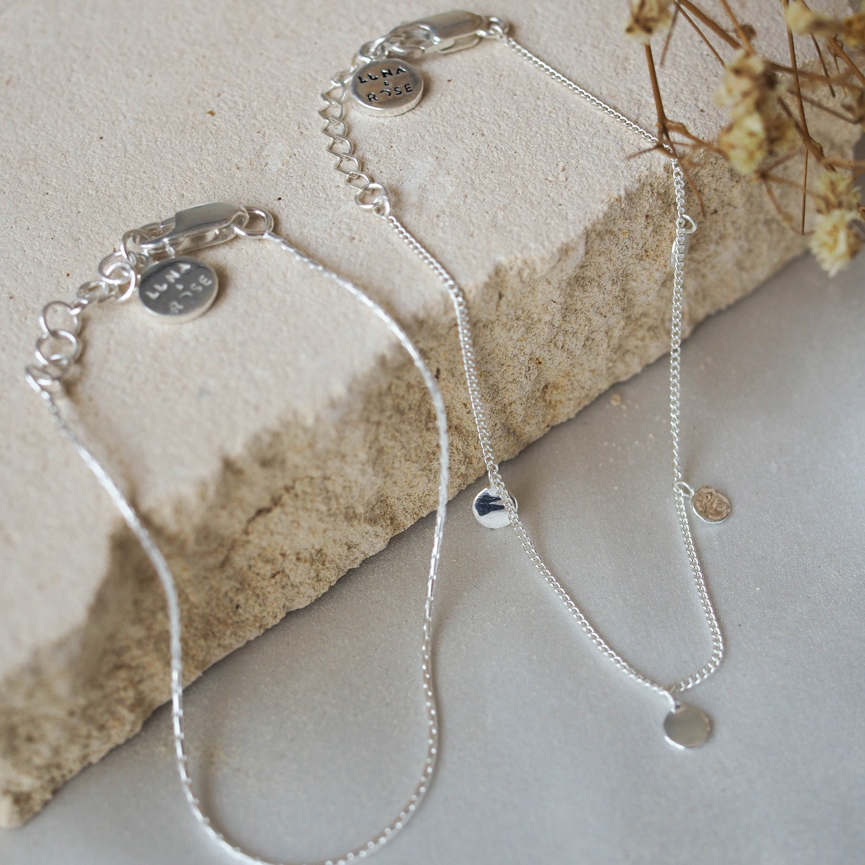 Two delicate silver chain bracelets with circular charms, displayed on beige stone with dried flowers.