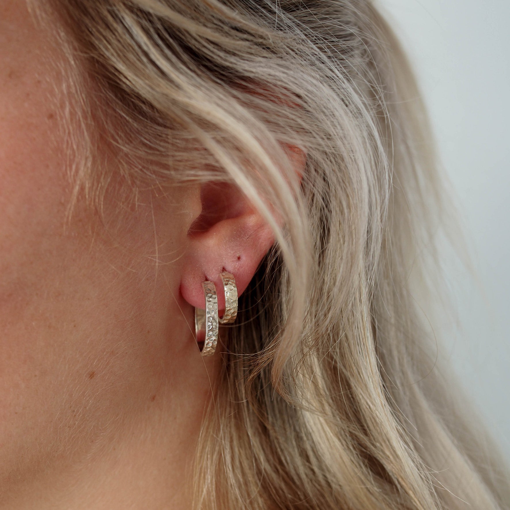 Close-up of two hammered silver hoop earrings worn on an ear with blonde hair.