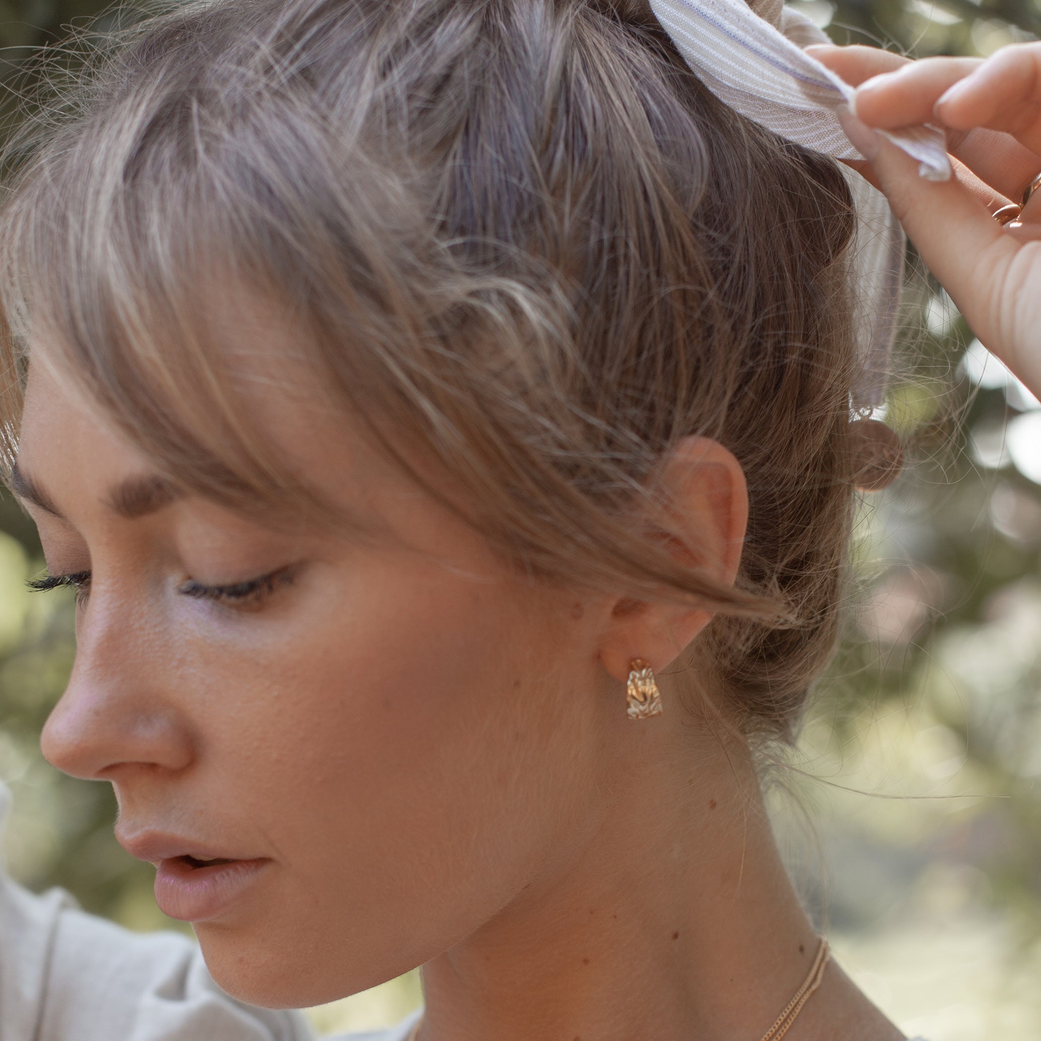 A person adjusting their hairband while wearing a gold textured hoop earring.