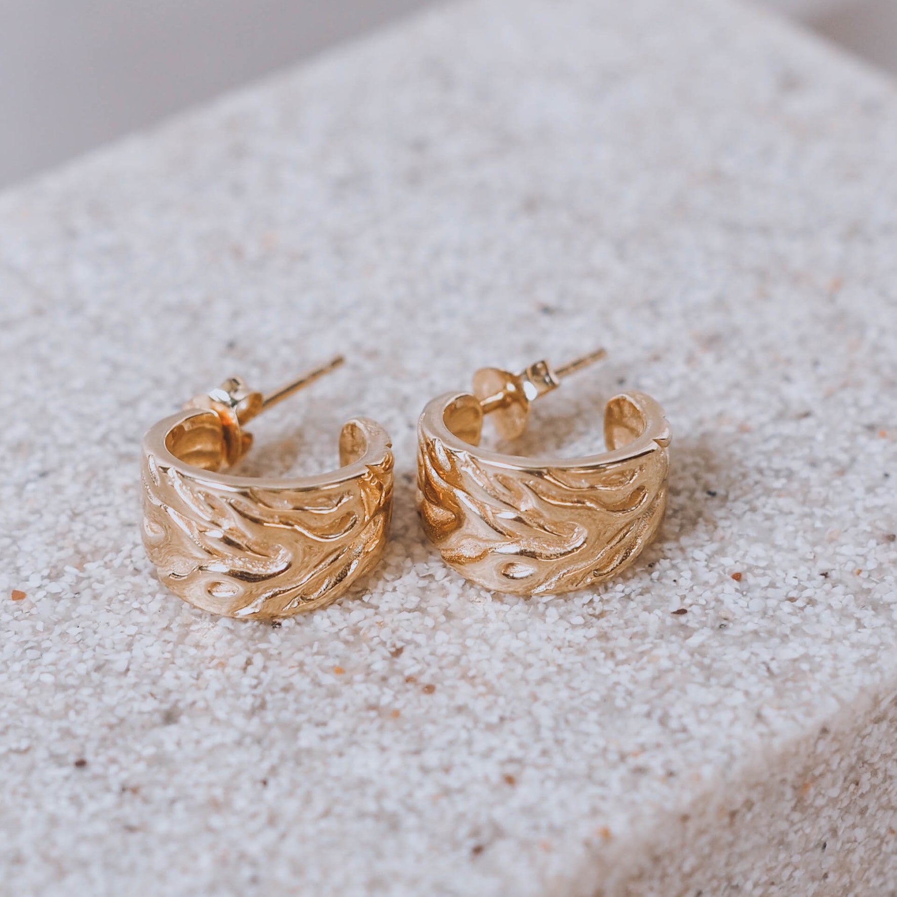 Detailed close-up of gold textured hoop earrings on a sandy surface.