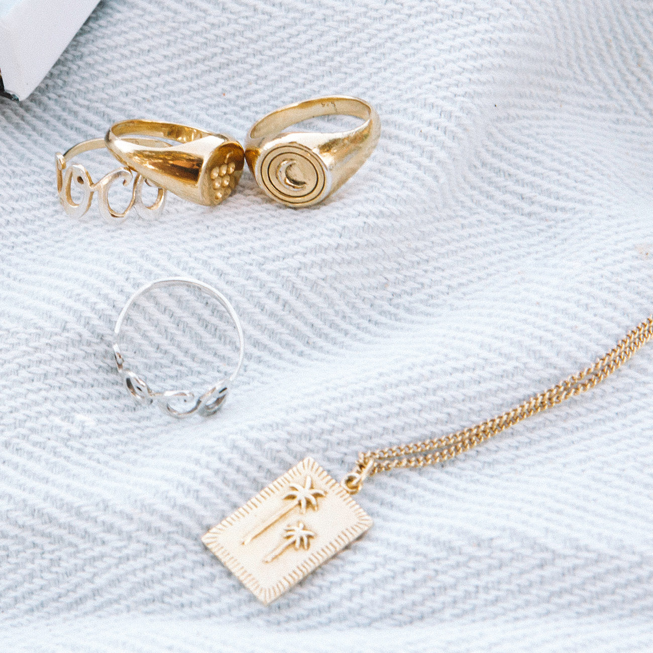 A collection of jewelry placed on a white textured cloth, including a gold rectangular pendant necklace with two raised palm tree designs and several gold rings.