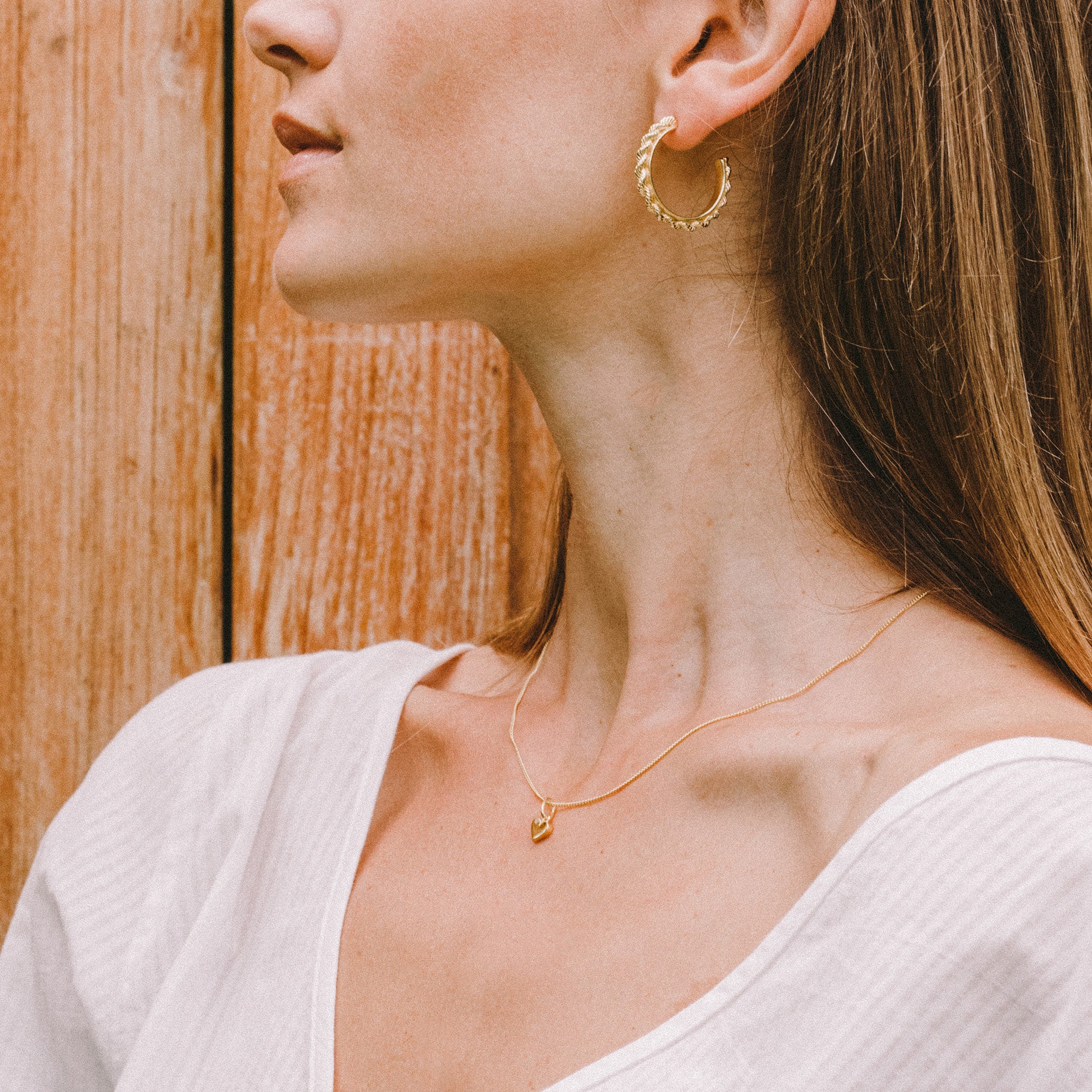A person wearing a gold hoop earring and a gold necklace with a small heart-shaped pendant, set against a wooden background.