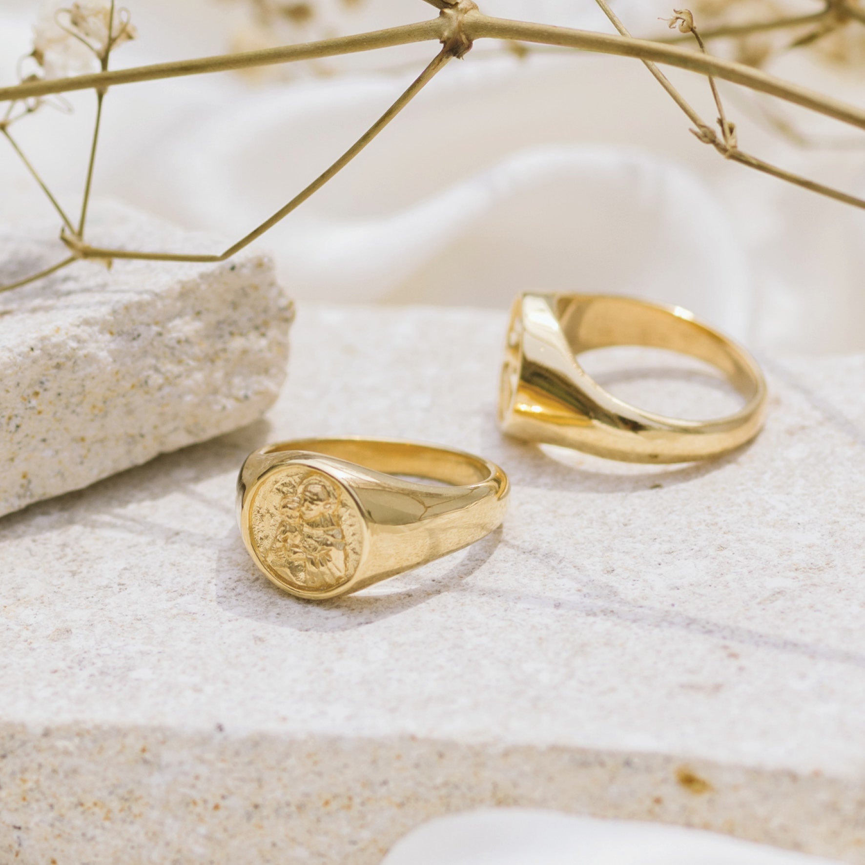 Two gold signet rings with round faces featuring a raised religious figure design, displayed on a light stone surface with some dried plants in the background.