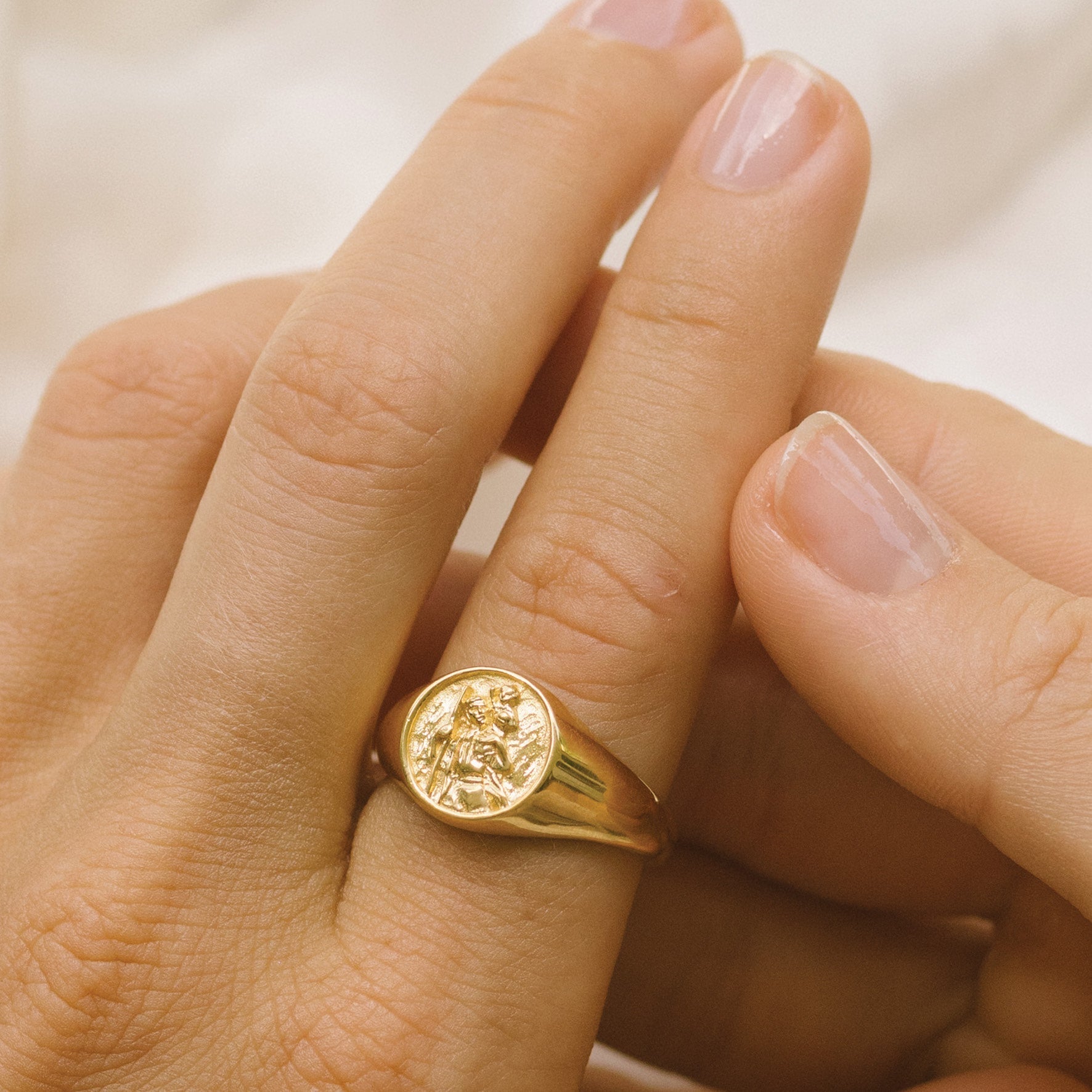 A close-up of a hand wearing a gold signet ring with a round face featuring a raised religious figure design, against a soft background.