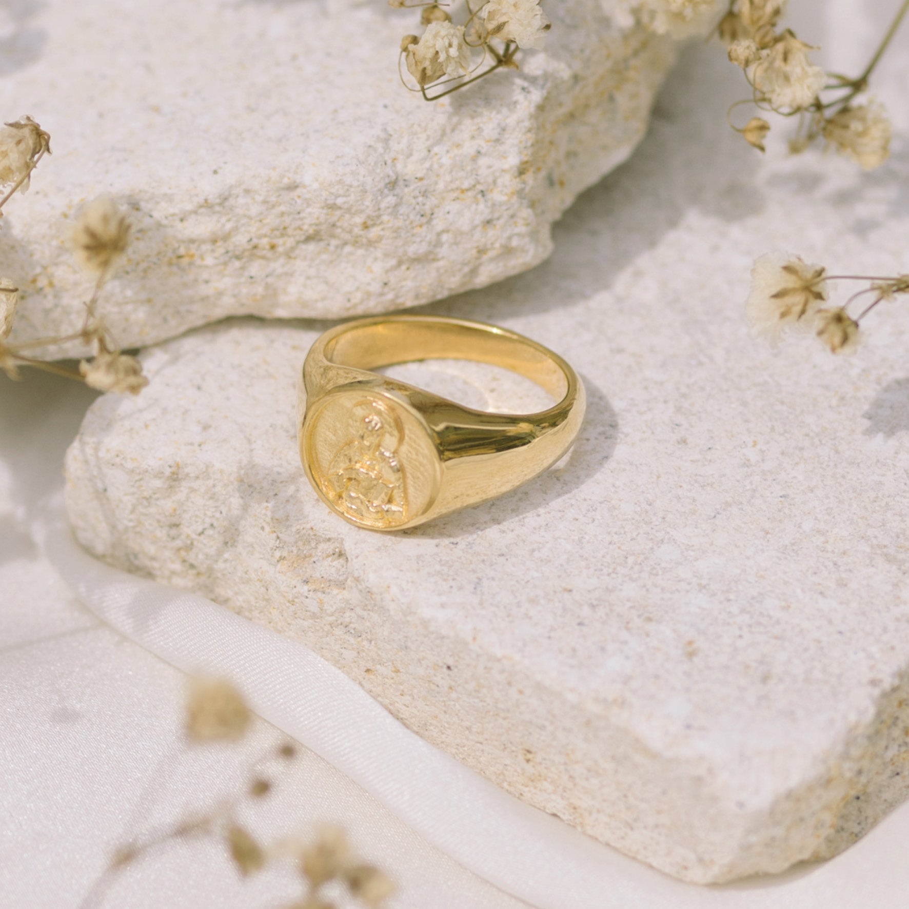 A gold signet ring with round faces featuring a raised religious figure design, displayed on a light stone surface with some dried plants in the background.