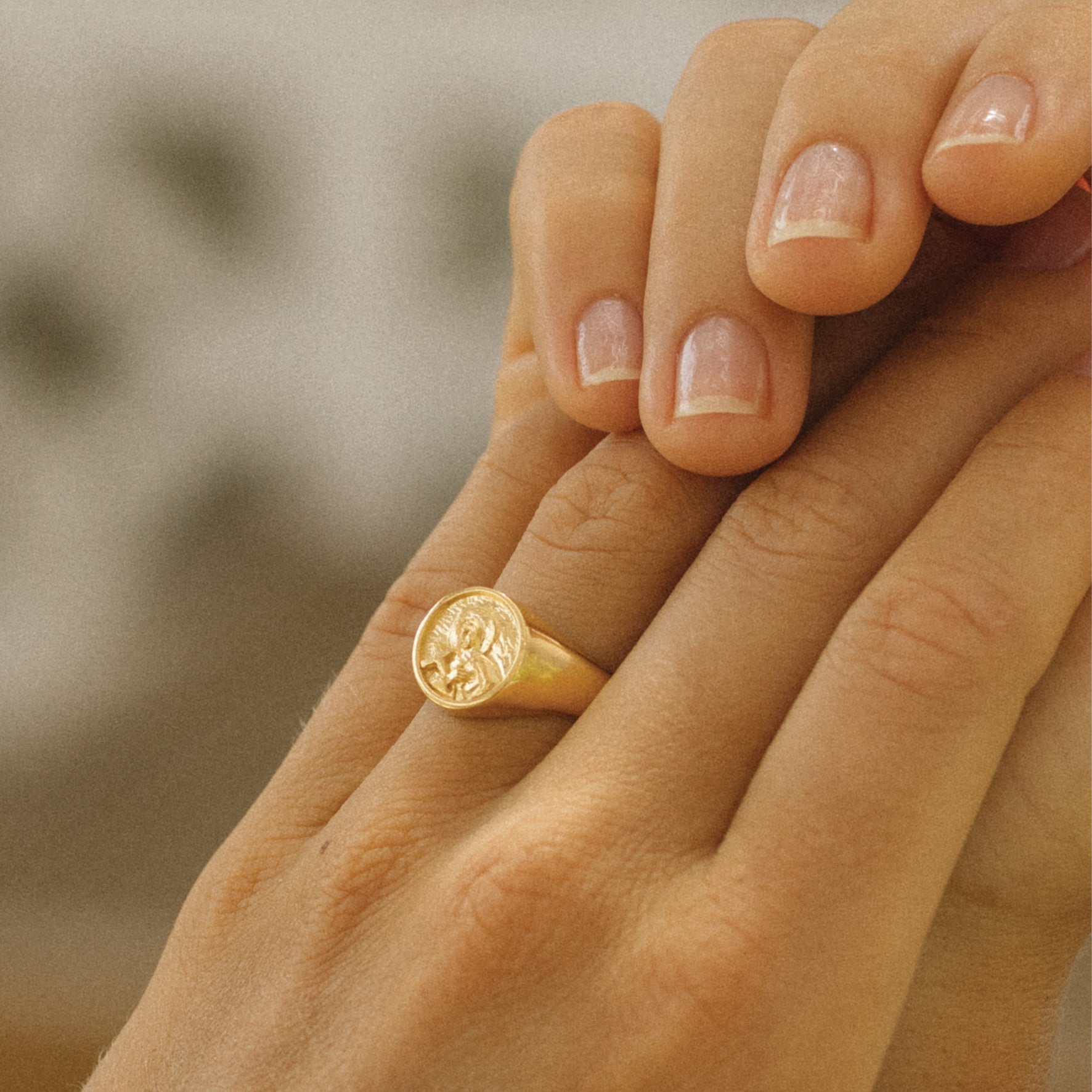 Close-up of a hand wearing a gold signet ring with a round face featuring a raised religious figure design, against a soft background.
