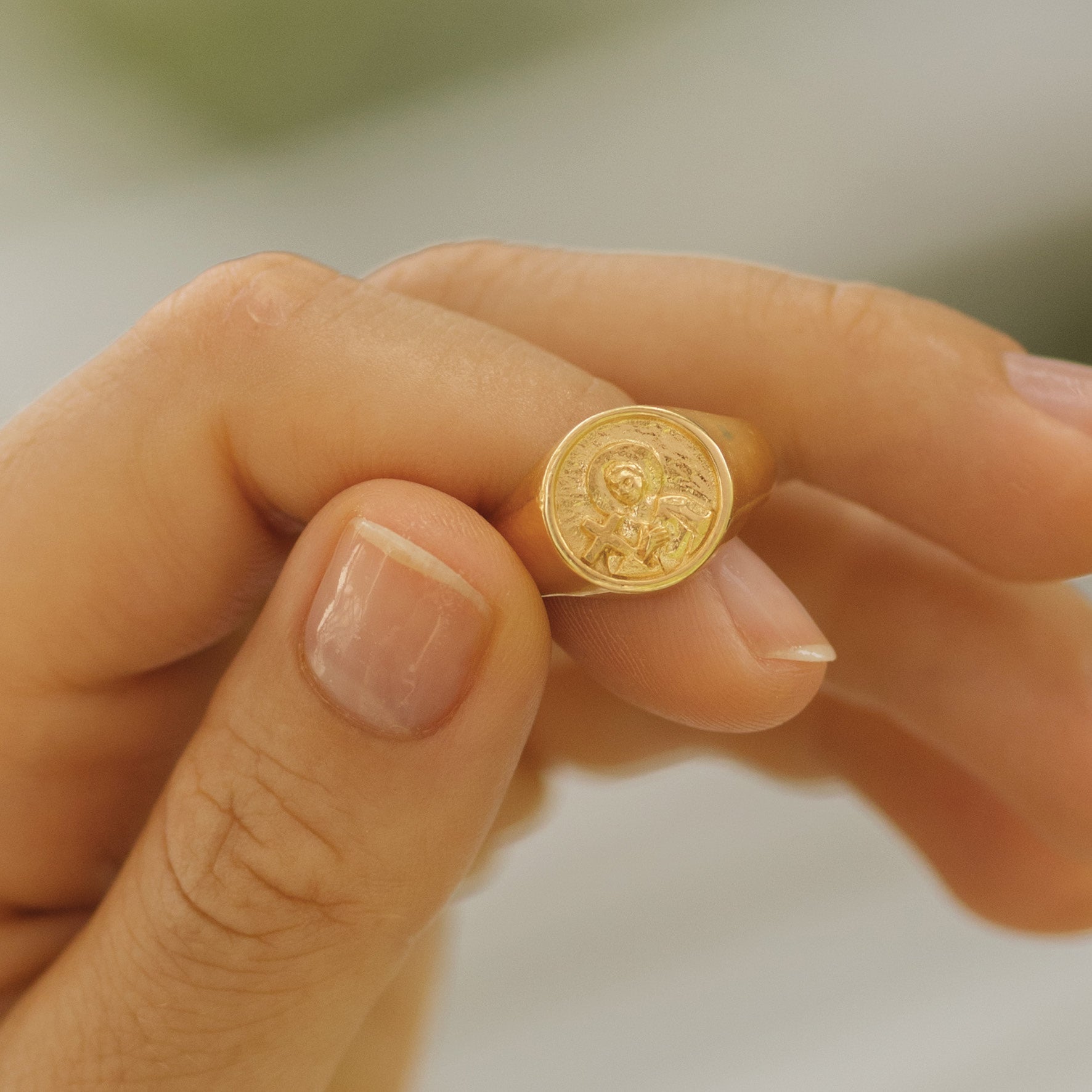Close-up of a hand holding a gold signet ring with a round face featuring a raised religious figure design, against a soft background.