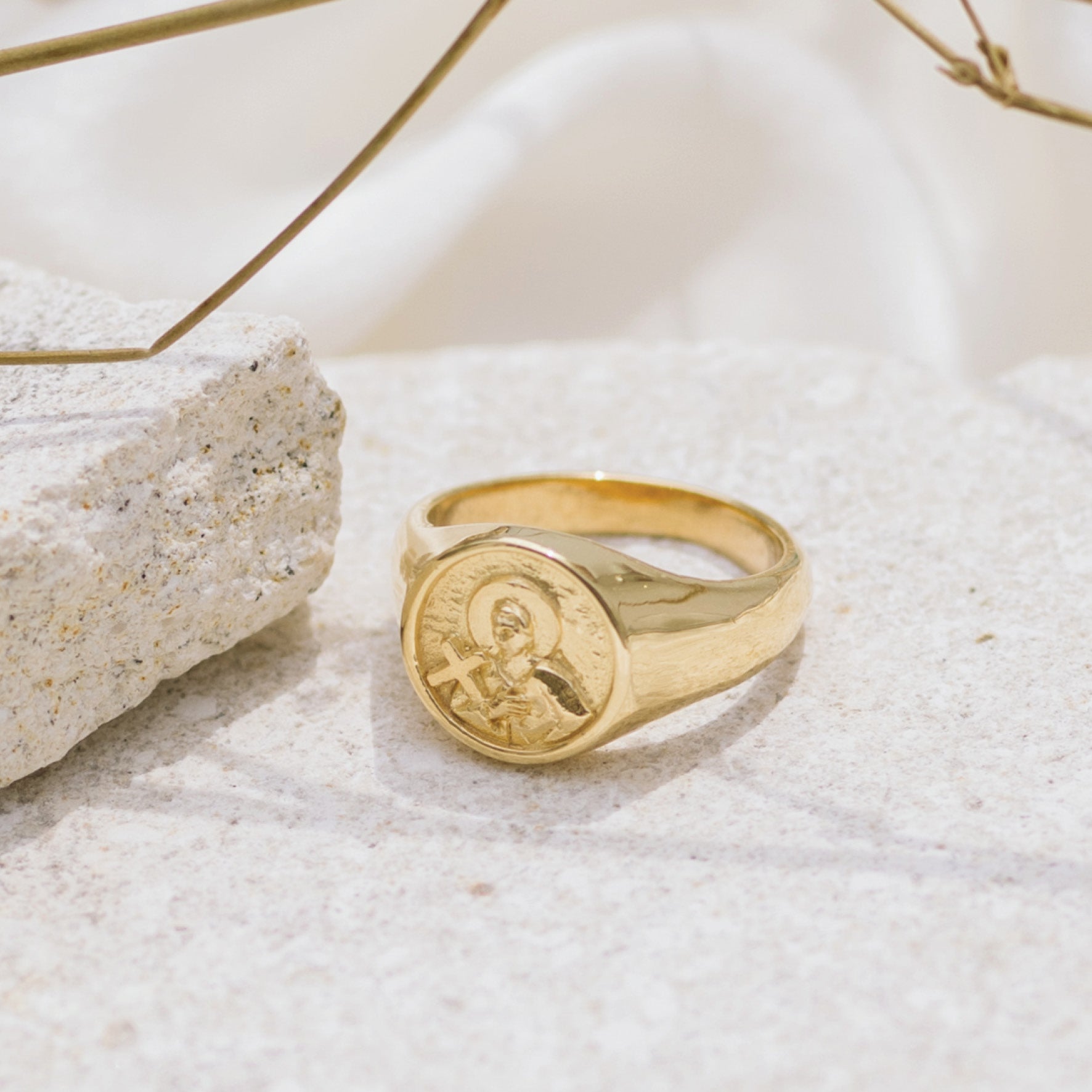 A gold signet ring with round faces featuring a raised religious figure design, displayed on a light stone surface with some dried plants in the background.