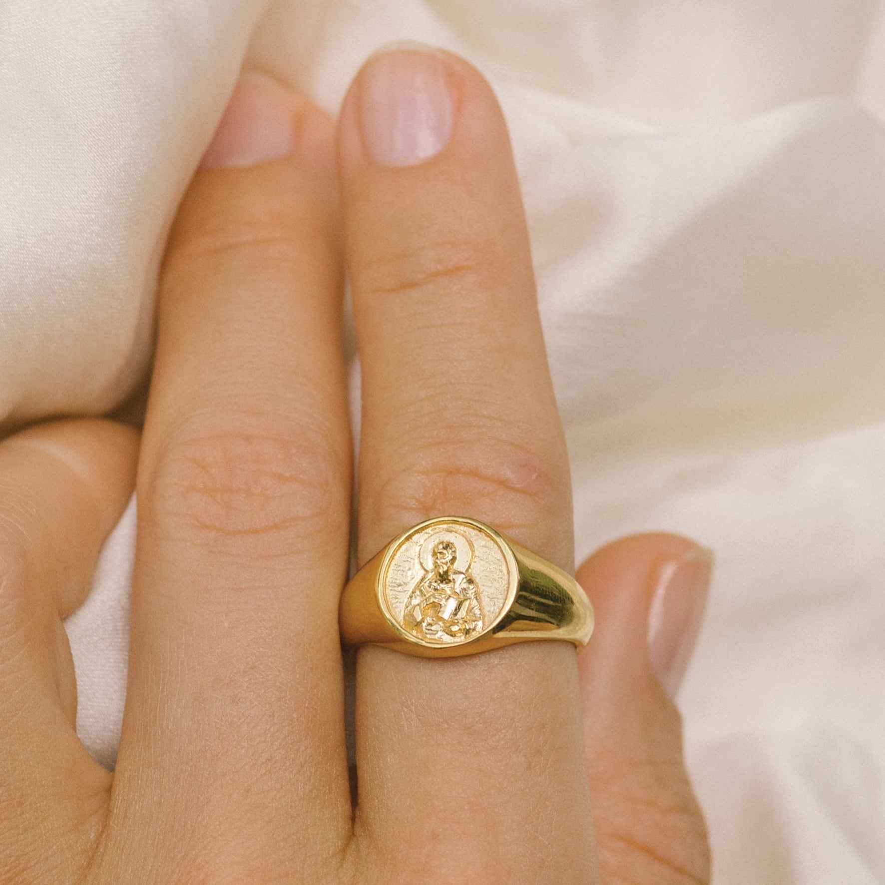 Close-up of a hand wearing a gold signet ring with a round face featuring a raised religious figure design, against a soft background.