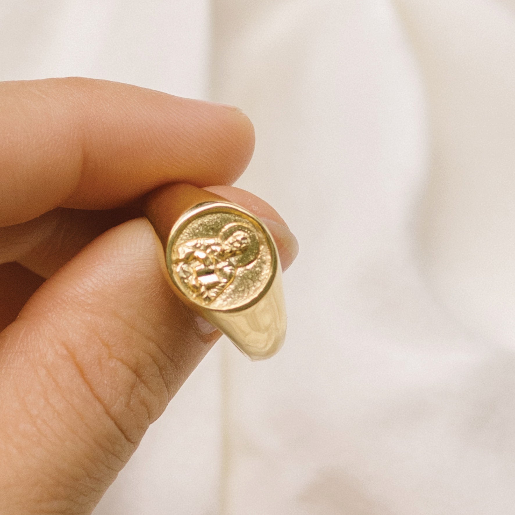 Close-up of a hand holding a gold signet ring with a round face featuring a raised religious figure design, against a soft background.
