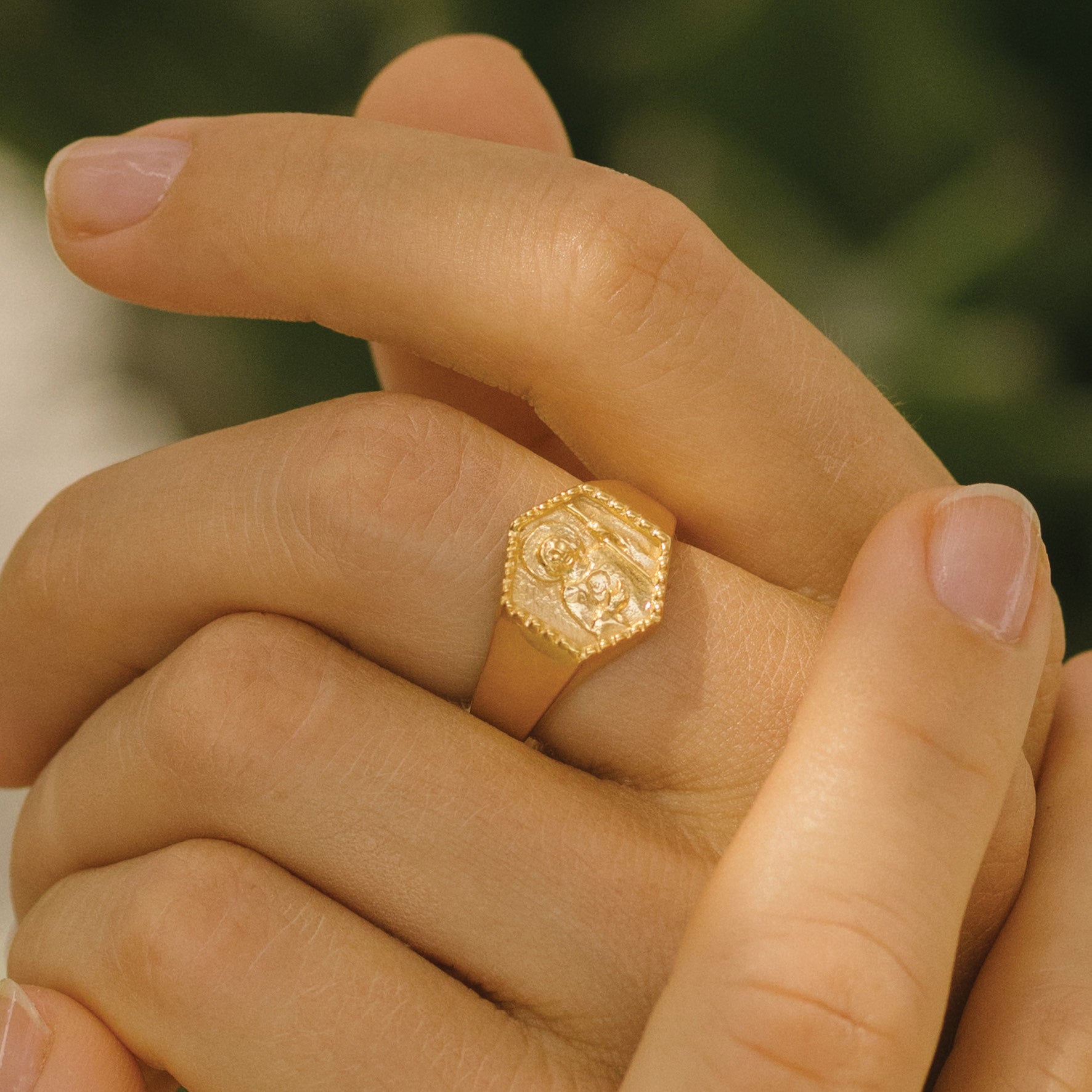 Close-up of a hand wearing a gold signet ring featuring a raised religious figure design, against a green background.