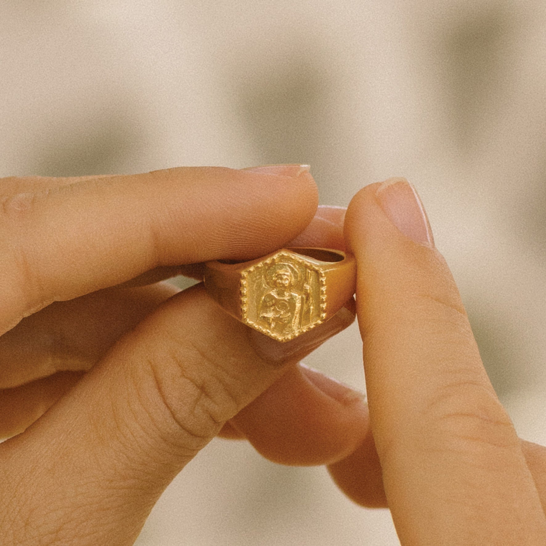Close-up of a hand holding a gold signet ring featuring a raised religious figure design, against a soft background.