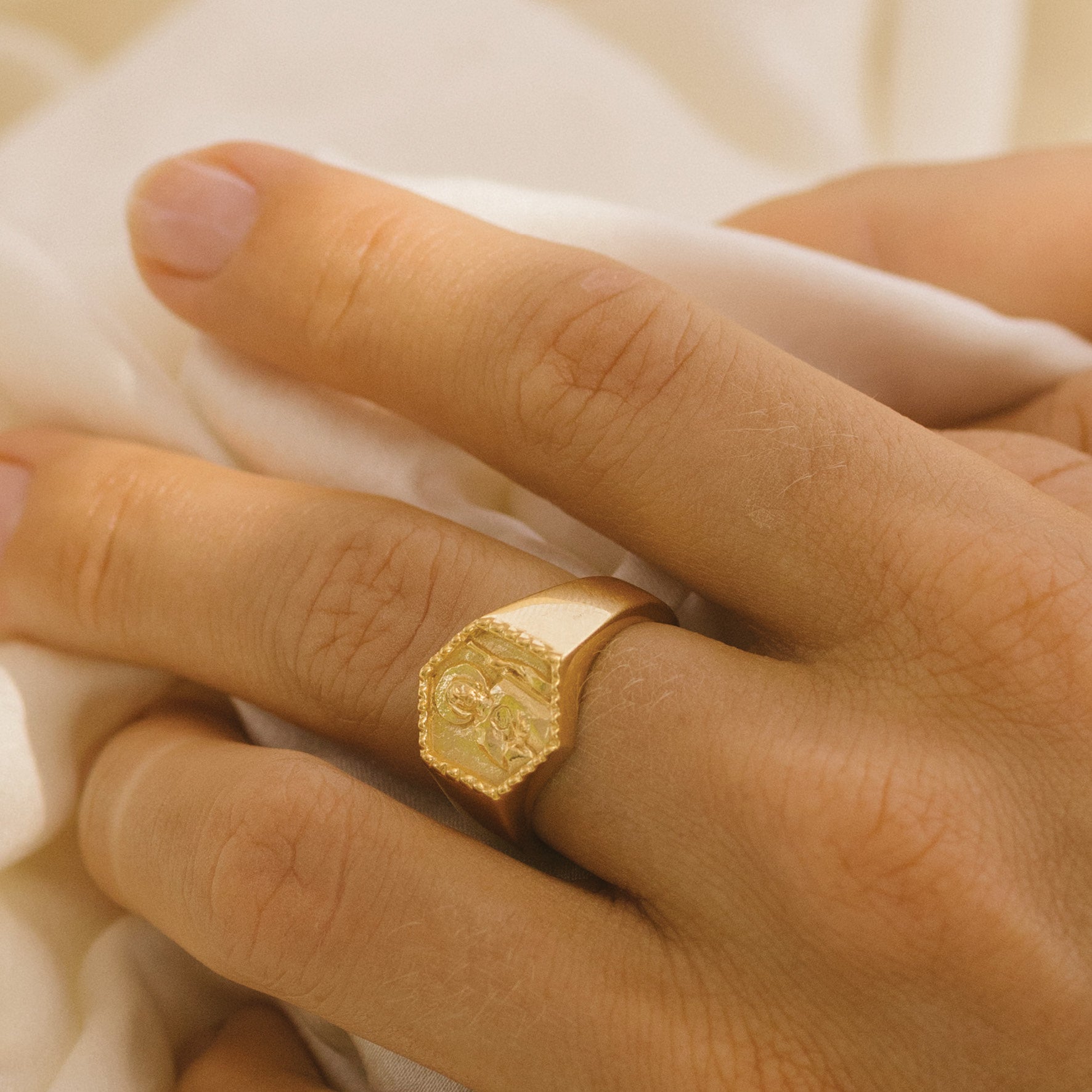 Close-up of a hand wearing a gold signet ring featuring a raised religious figure design, against a soft background.