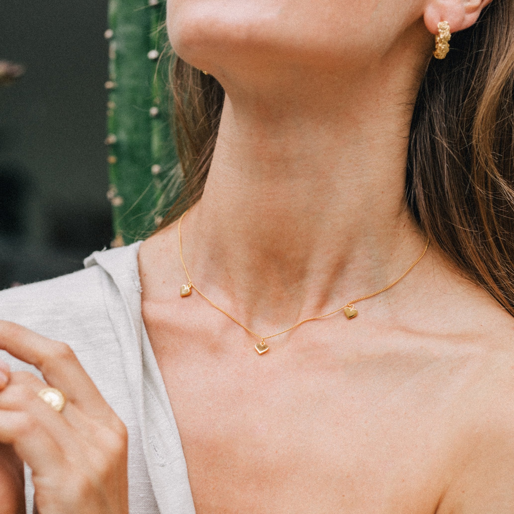 A close-up of a person wearing a gold necklace with three small heart pendants and gold earrings, set against a background with green elements.