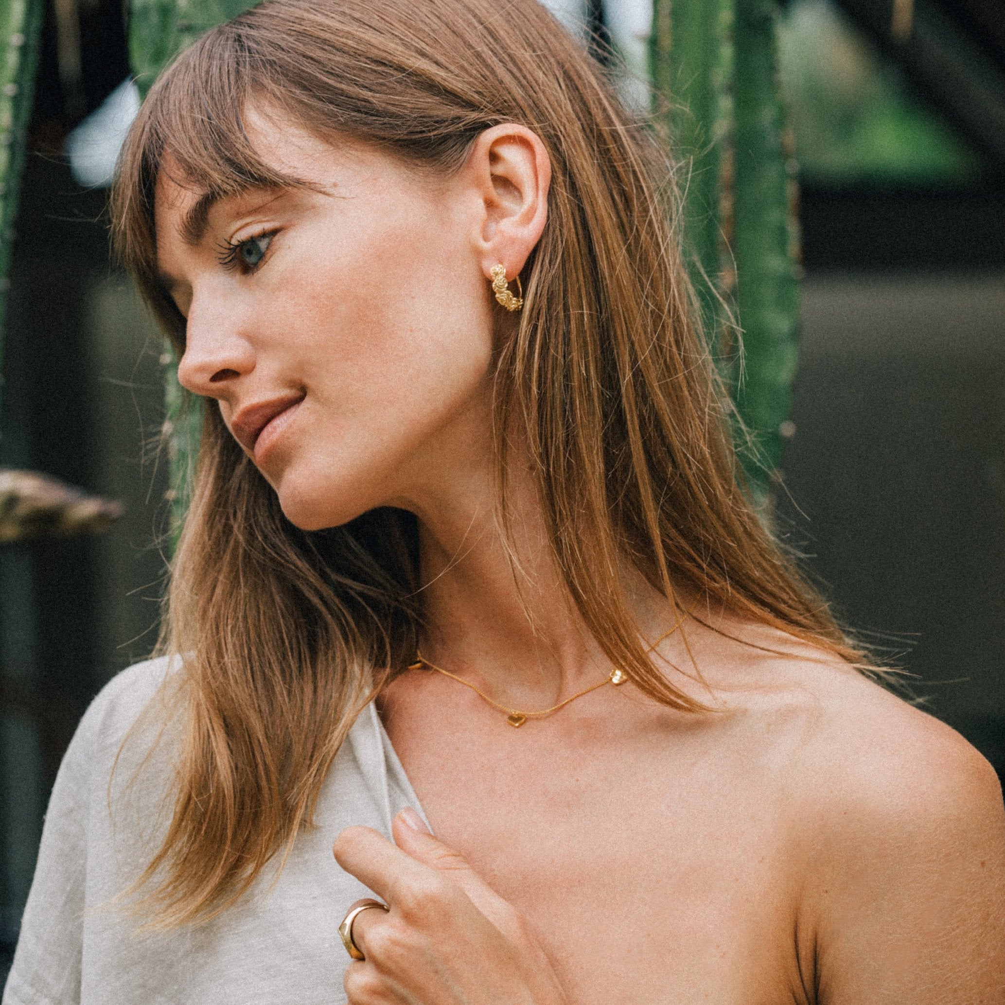 A person wearing a gold necklace with three small heart pendants and gold earrings, posing outdoors.