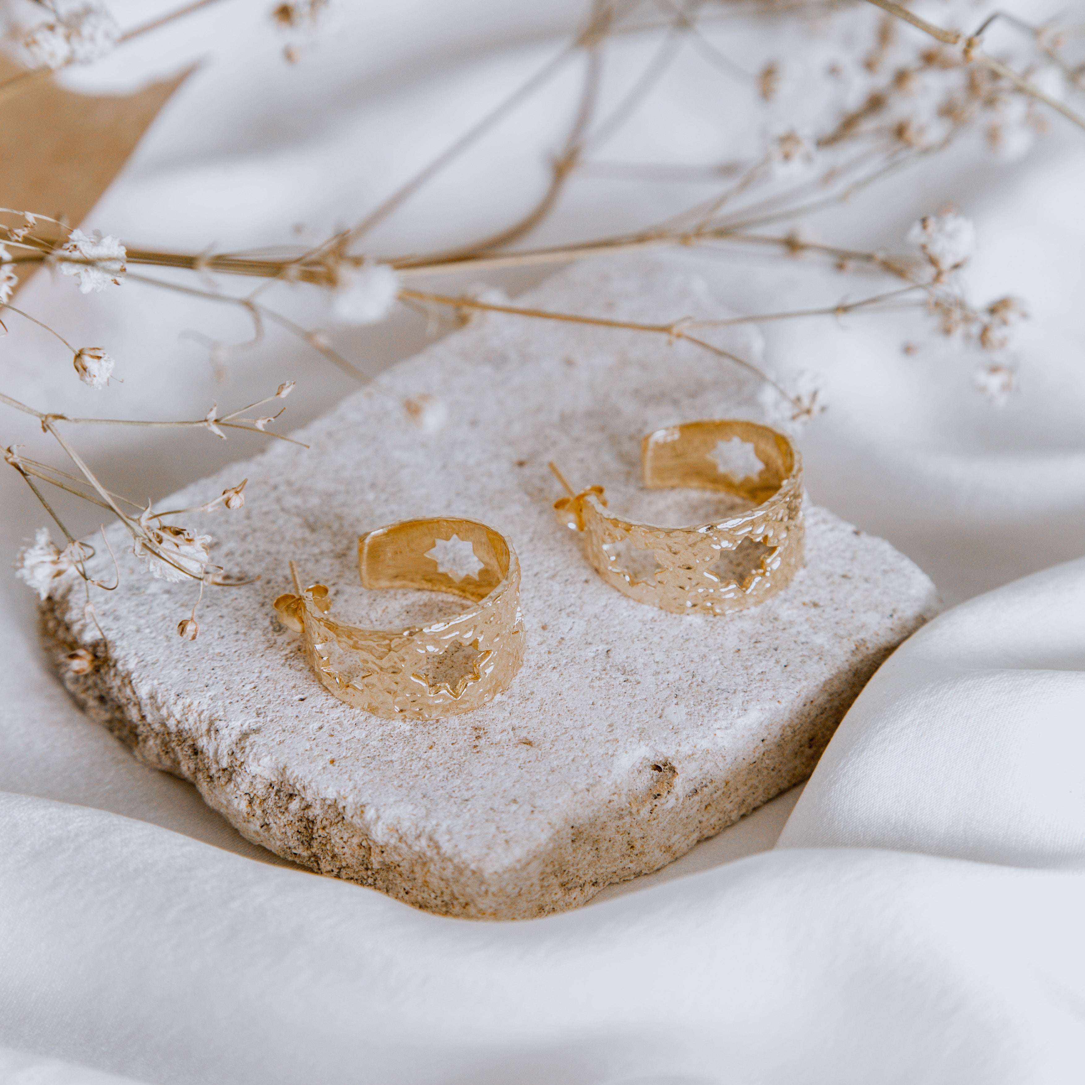 Golden earrings with hoops featuring stars, displayed on a light stone surface with some dried plants in the background.