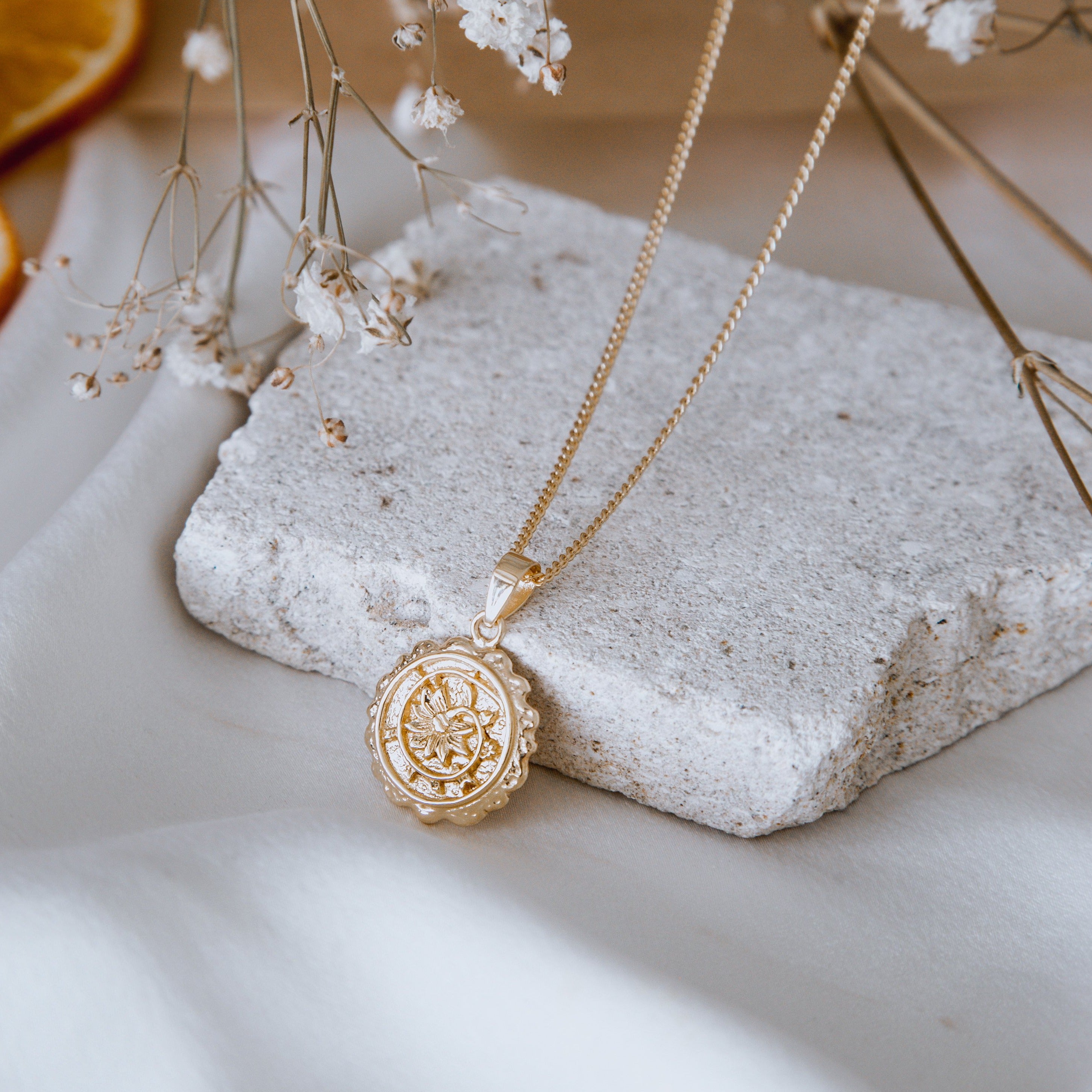 A gold necklace with an intricate round pendant is displayed on a textured stone, accompanied by delicate white flowers and soft, neutral-colored fabric.