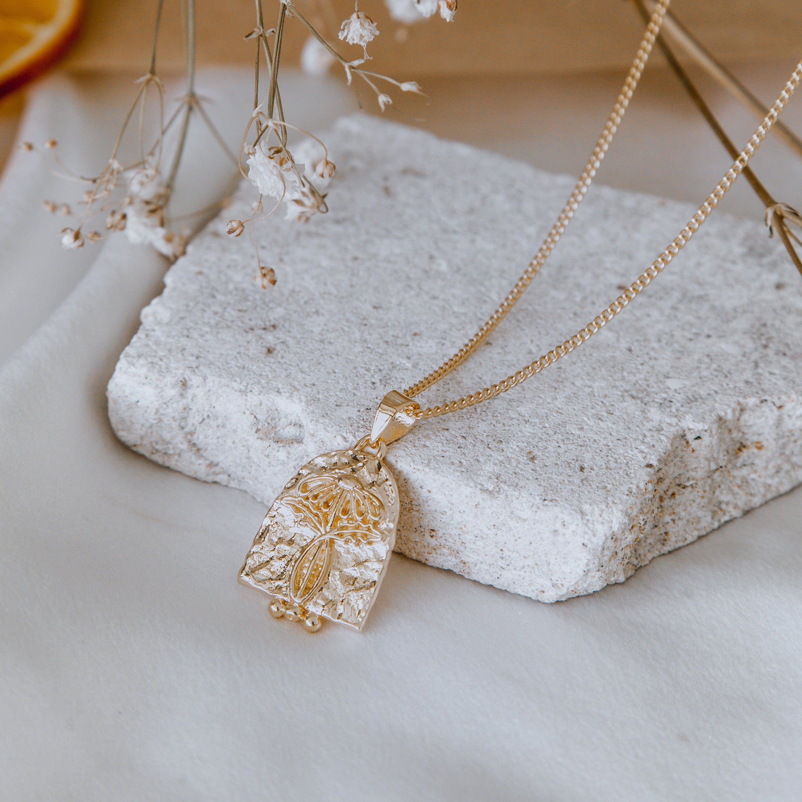 A close-up of a necklace displayed on a beige stone with delicate white flowers surrounding it.