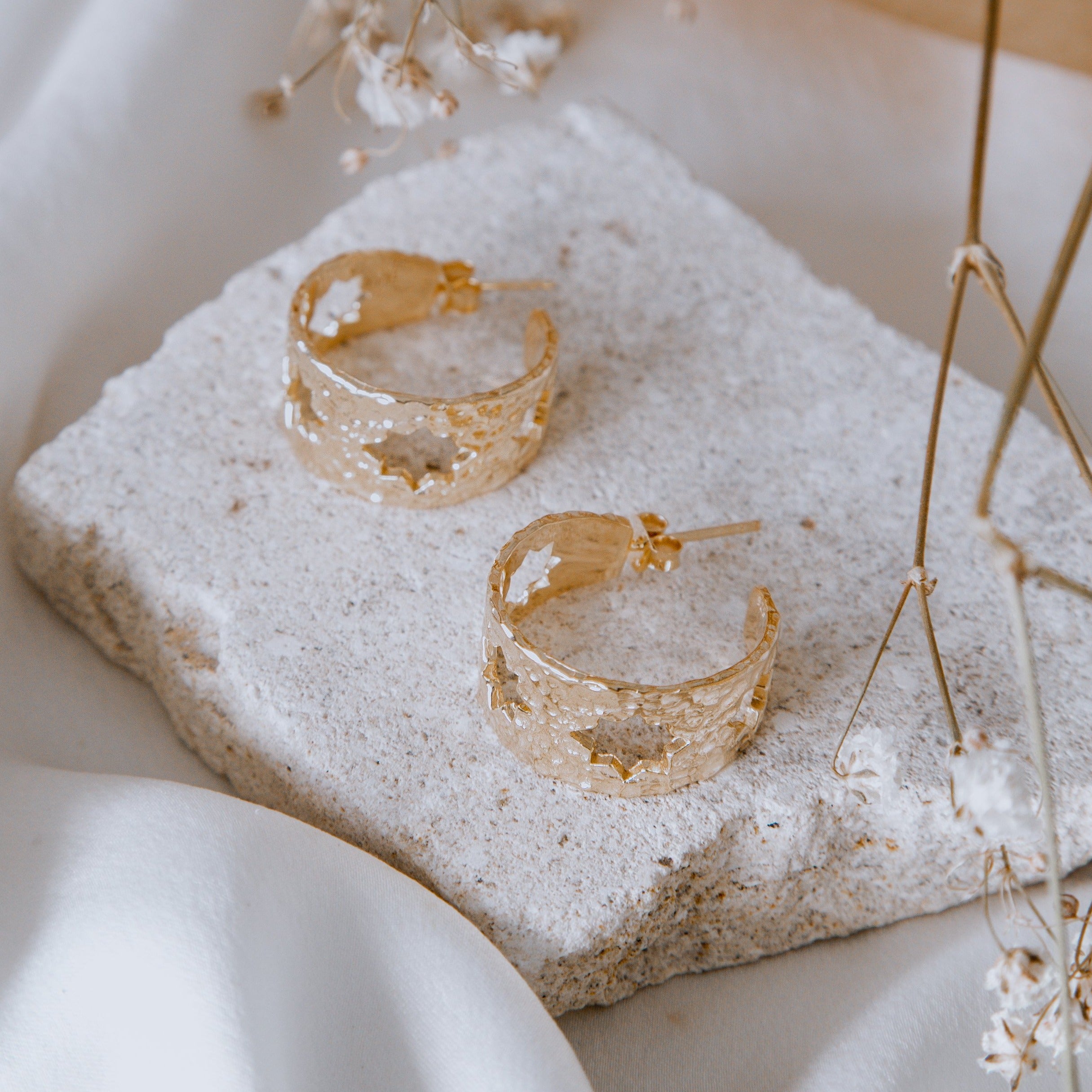 Golden earrings displayed on a beige stone with delicate white flowers surrounding it.