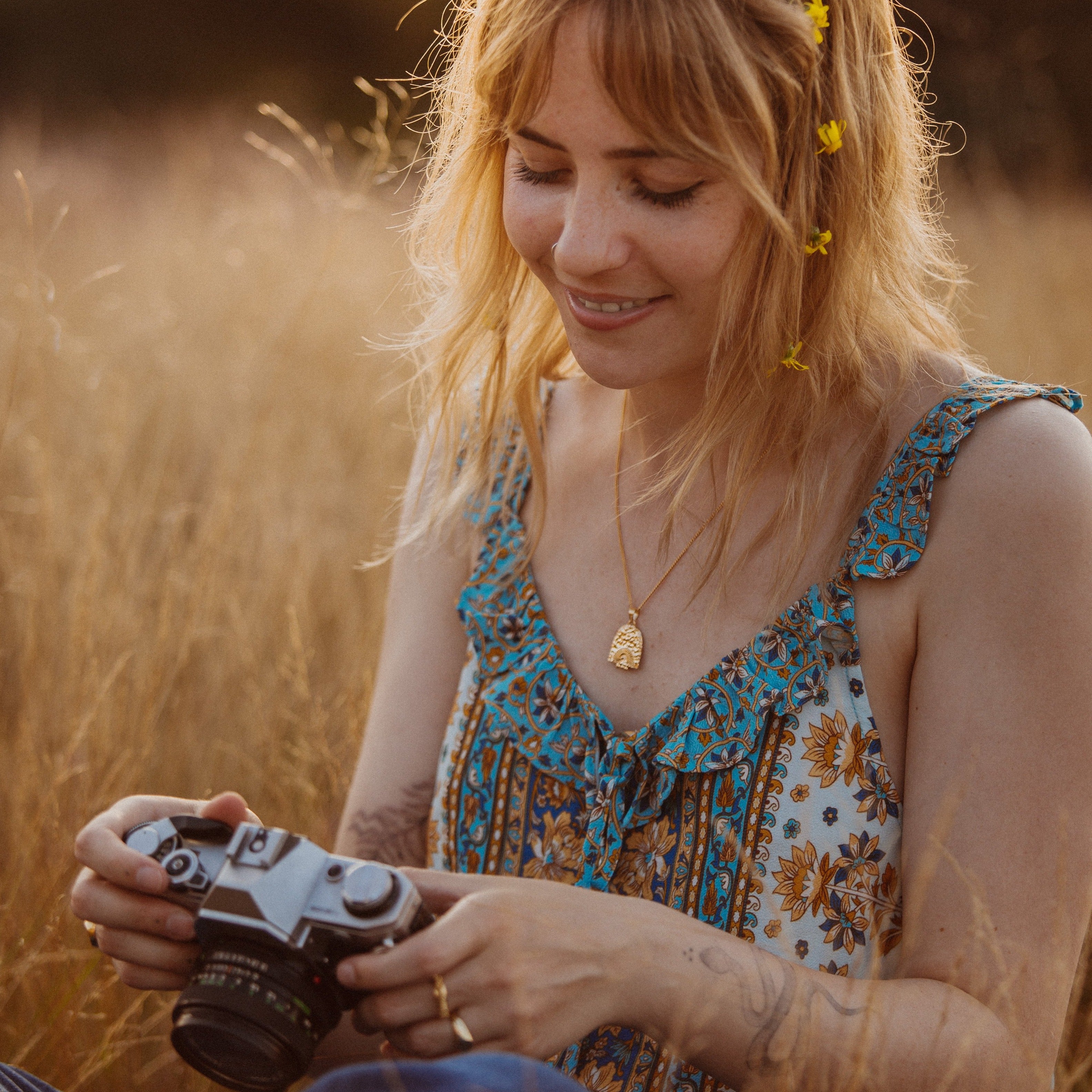A woman wearing a patterned dress and a gold necklace with a pendant sits in a field of tall grass, holding a vintage camera and smiling.