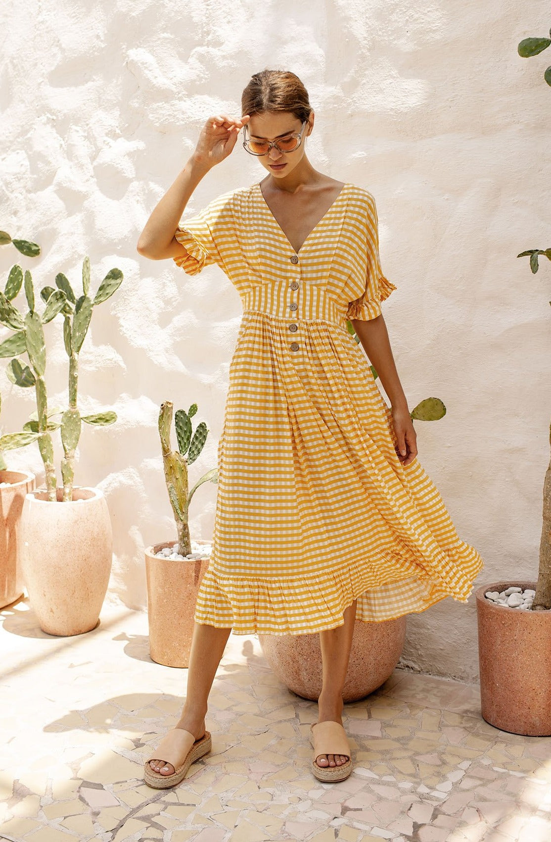 A woman wearing a yellow and white gingham dress with a V-neckline and buttons down the front, accessorized with sunglasses and tan platform sandals, standing among potted plants.