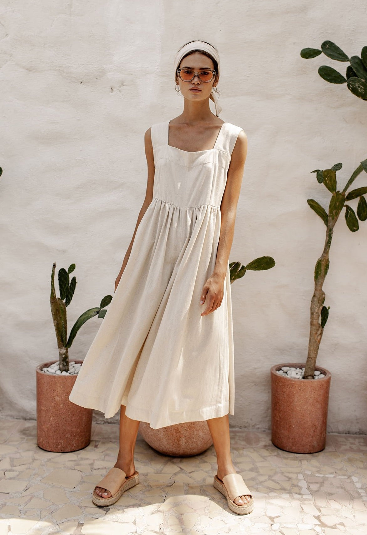 A woman wearing a sleeveless beige dress and tan sandals, accessorized with a headband and sunglasses, stands in front of potted plants.