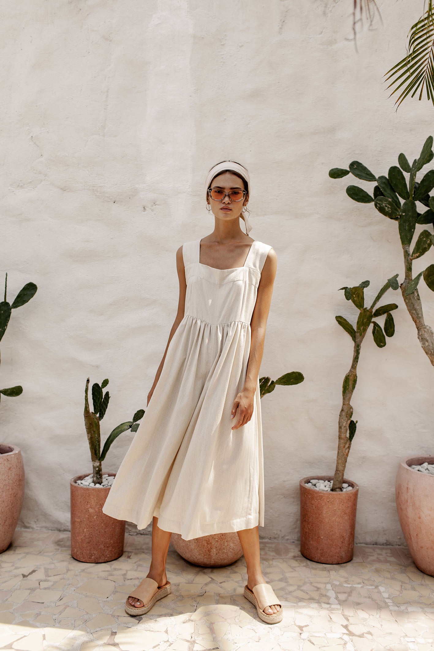 A woman wearing a sleeveless beige dress and tan sandals, accessorized with a headband and sunglasses, stands in front of potted plants.