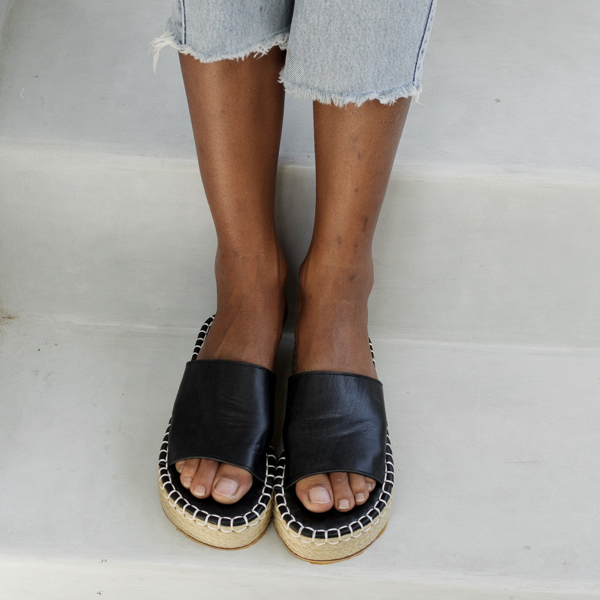 A close-up of a person wearing black slider sandals with a woven sole, paired with frayed hem jeans.