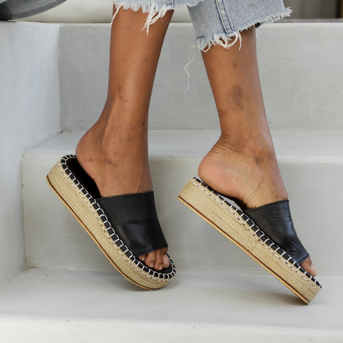 A side view of a person wearing black slider sandals with a woven sole, stepping on white stairs.