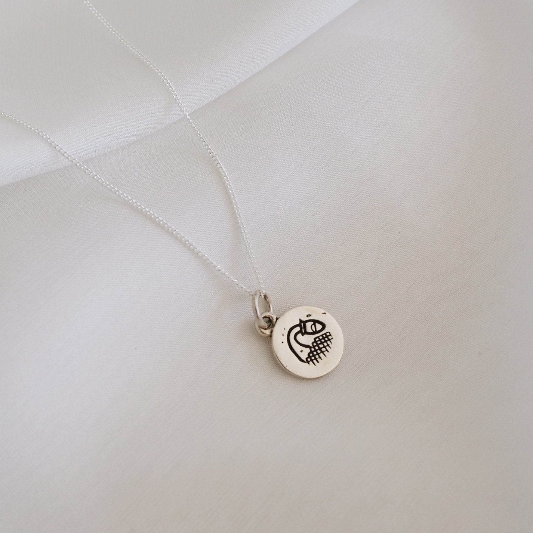 A simple silver necklace featuring a round pendant with a minimalist geometric design, displayed on a white fabric backdrop.