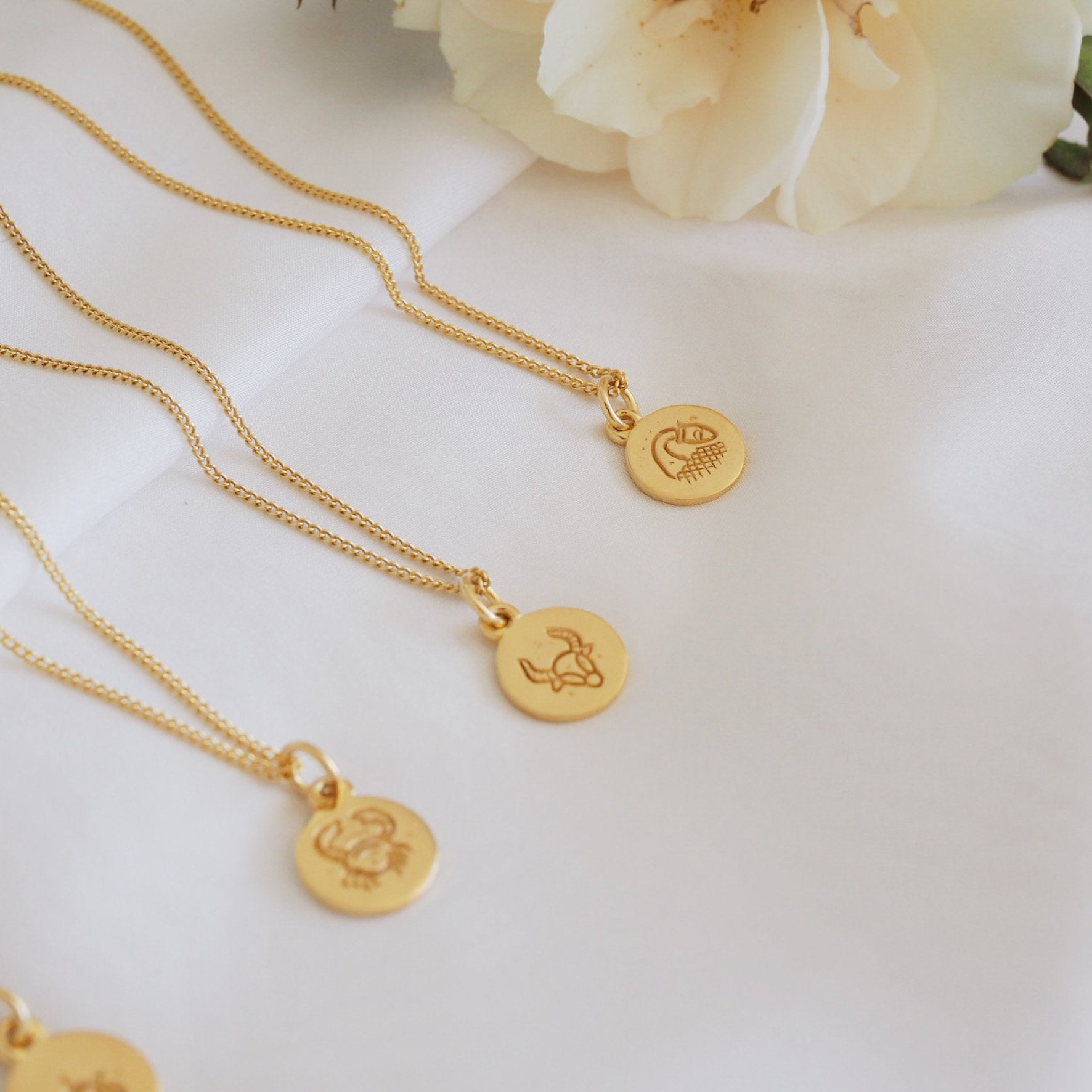Multiple gold necklaces featuring a round pendant with a minimalist geometric design, displayed on a white fabric backdrop.