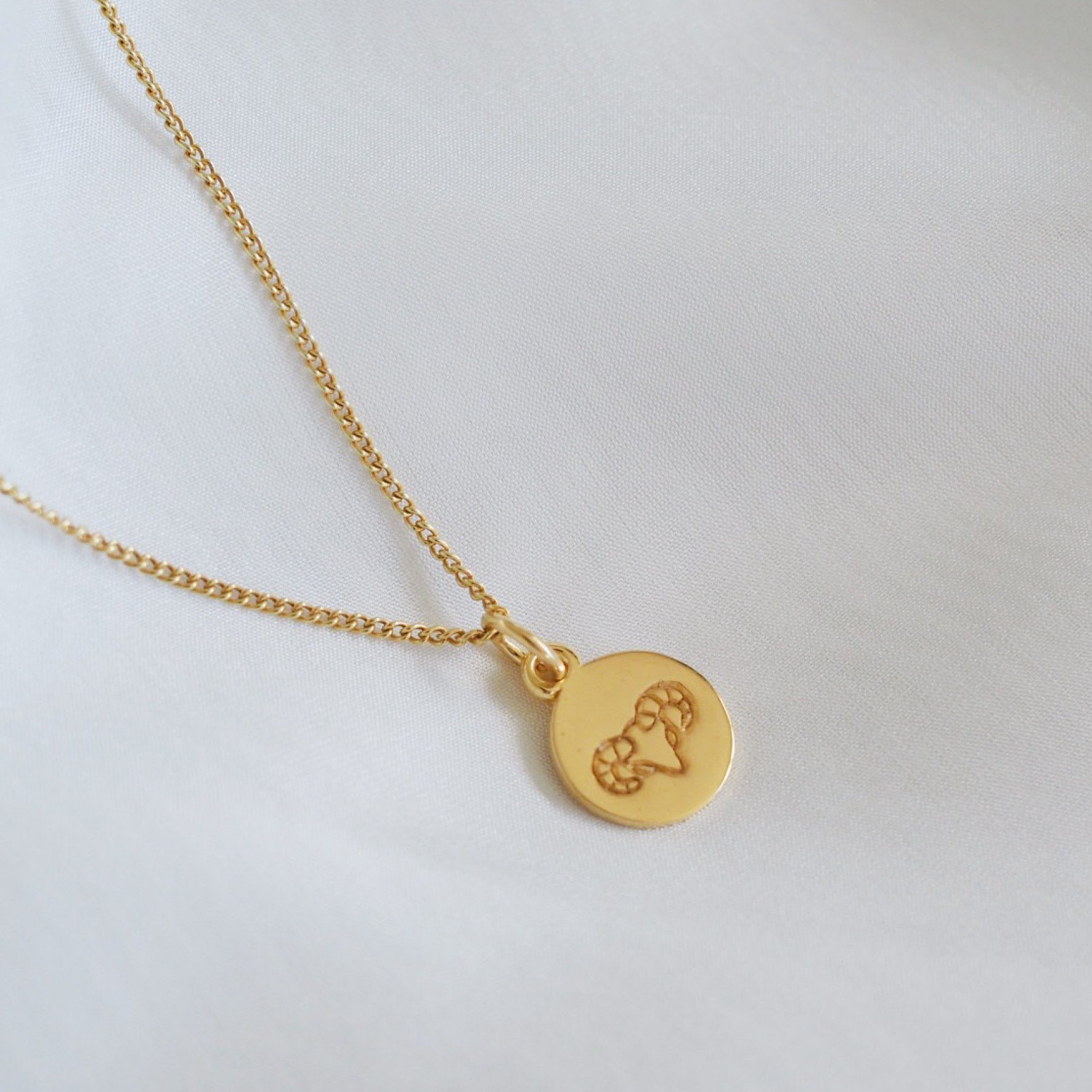 A simple gold necklace featuring a round pendant with a minimalist geometric design, displayed on a white fabric backdrop.