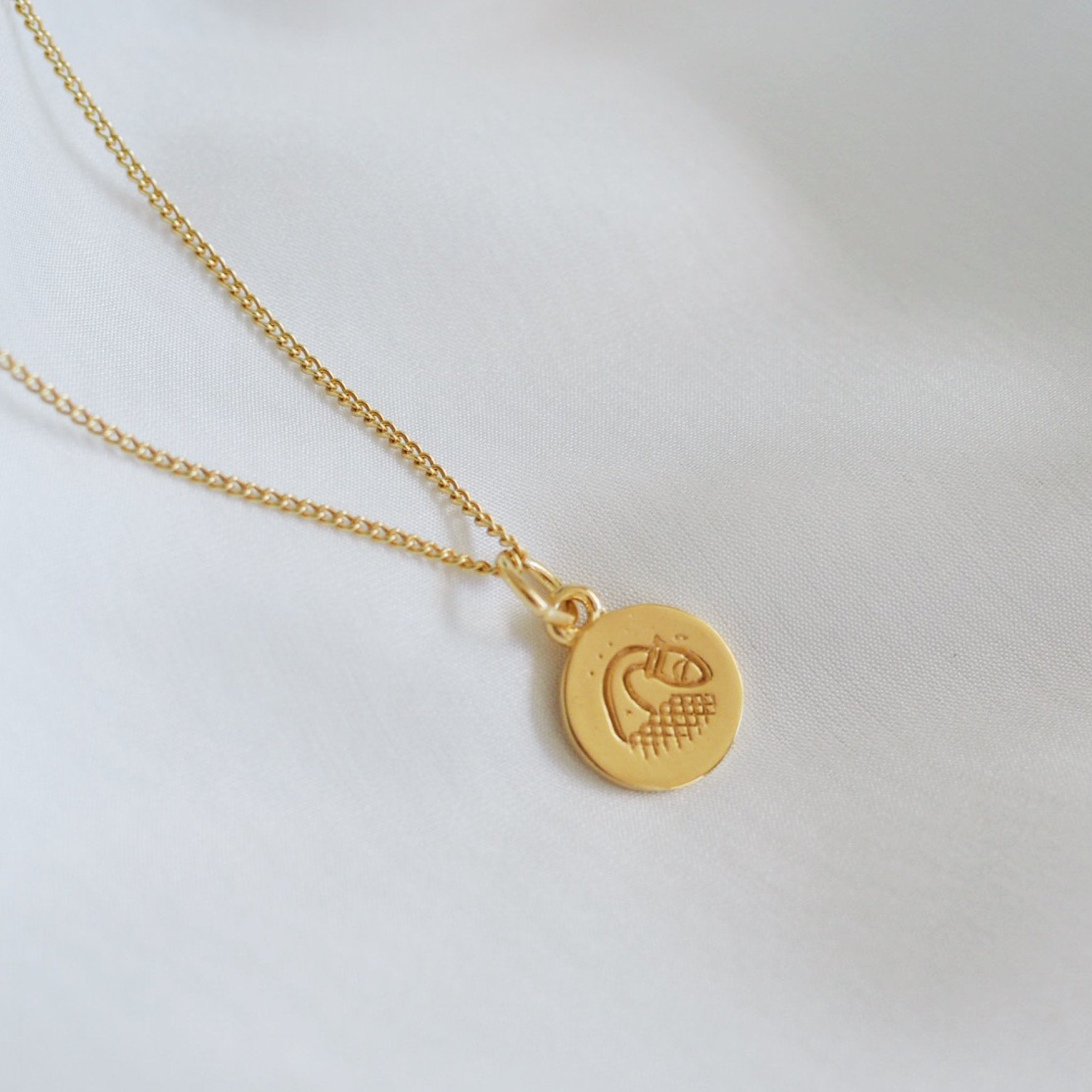 A simple gold necklace featuring a round pendant with a minimalist geometric design, displayed on a white fabric backdrop.