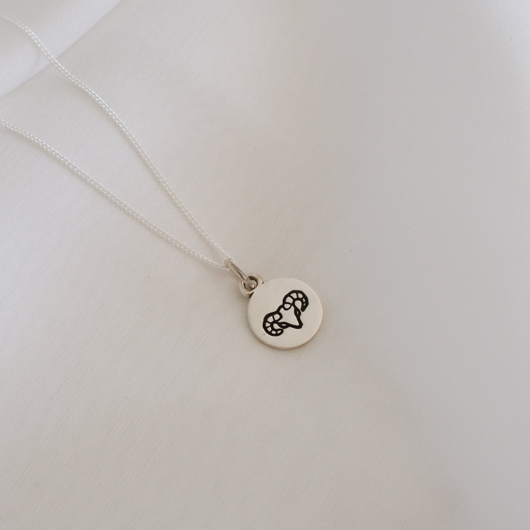 A simple silver necklace featuring a round pendant with a minimalist geometric design, displayed on a white fabric backdrop.
