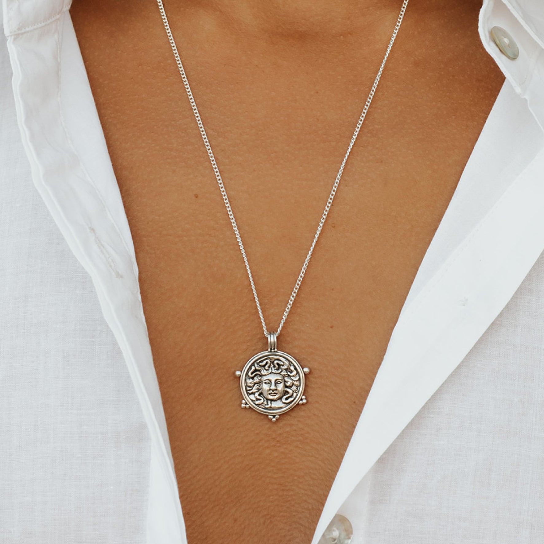 A silver Medusa pendant on a chain worn with a white shirt with an open collar.